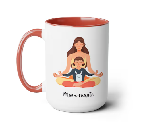 White mug with pink handle and rim, featuring an illustration of a mother and child meditating together, with the caption "Mom-maste".