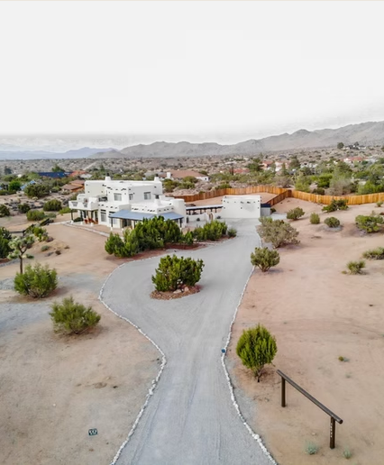 A modern white house with clean lines and flat roof, surrounded by desert landscape and sparse greenery, connecting via a curved gravel driveway, with mountains in the background.