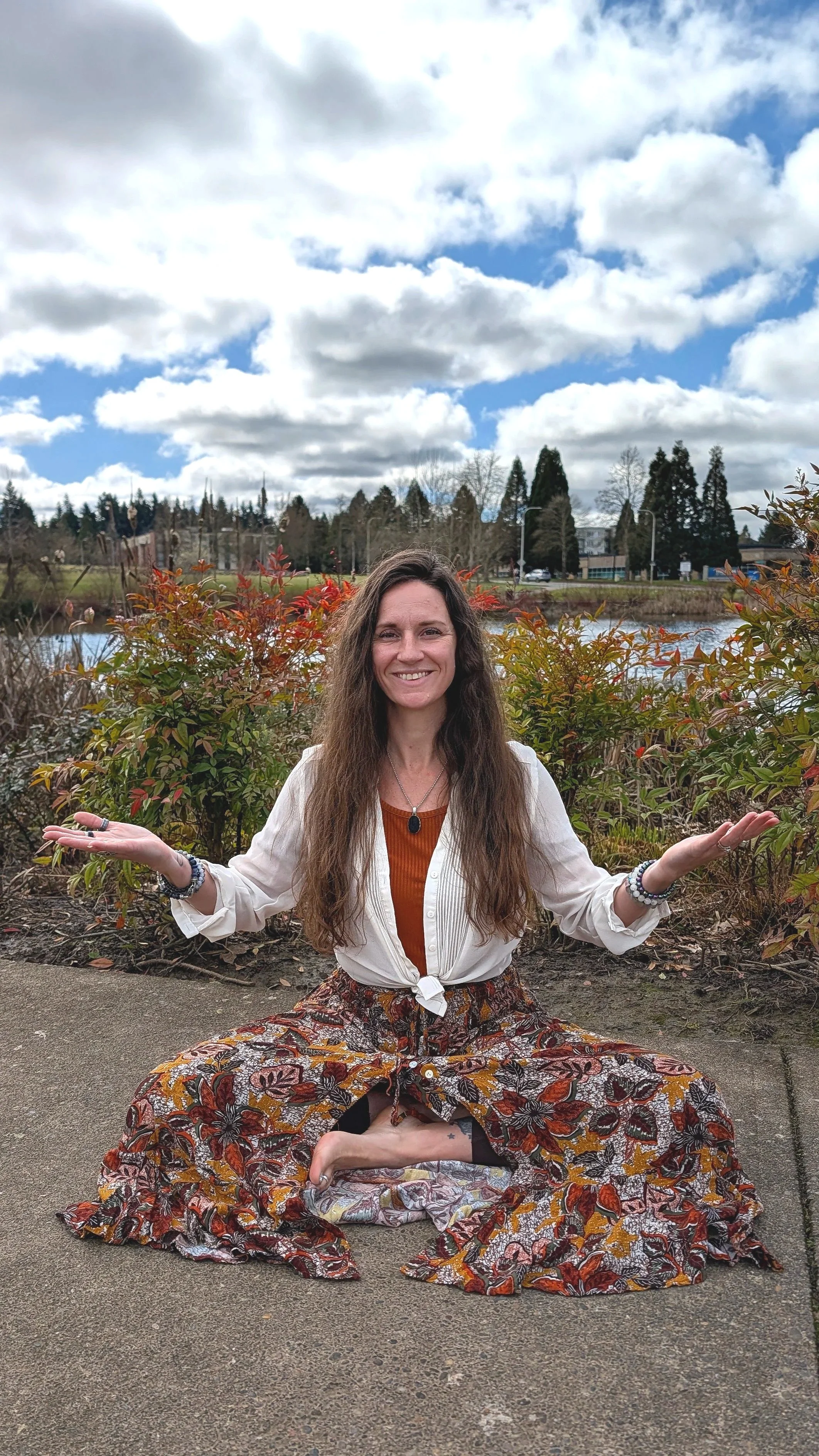 A woman with long wavy hair sitting cross-legged on the ground outdoors, smiling with her arms open, holding a small object in her left hand, against a backdrop of bushes, trees, a body of water, and a partly cloudy sky.