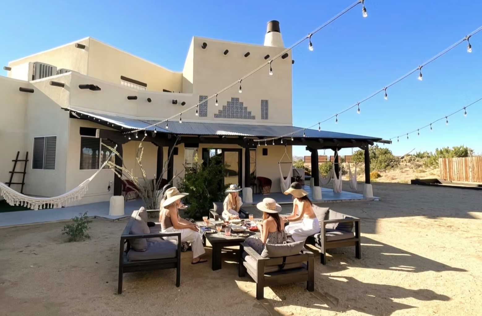 Group of women in sun hats enjoying a meal outdoors on a patio with sand ground, string lights overhead, and a modern house in the background.