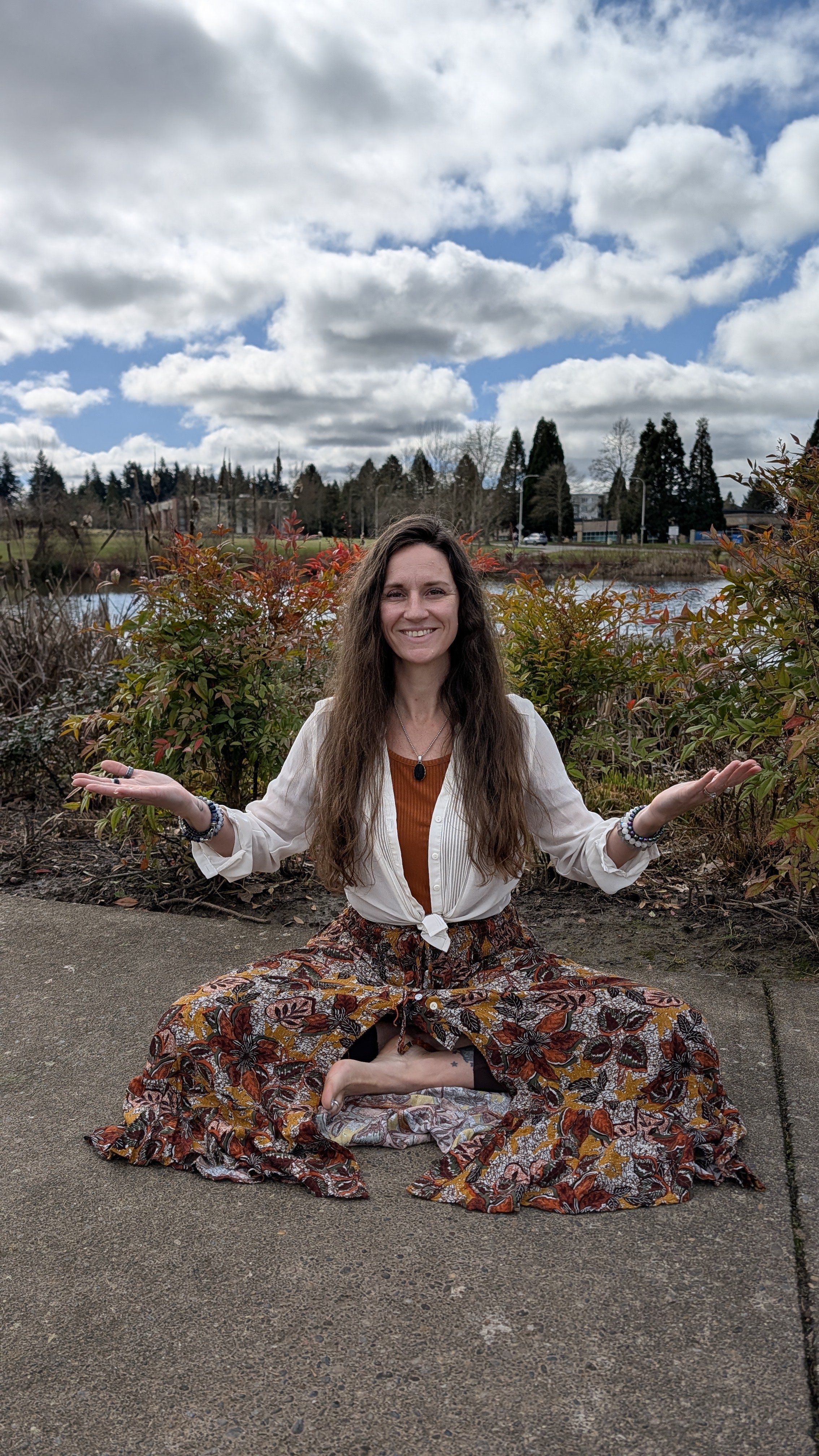 A woman practicing yoga or meditation outdoors near a pond or lake, sitting cross-legged on the ground in a peaceful pose with arms open and a smile, surrounded by plants and trees under a partly cloudy sky.