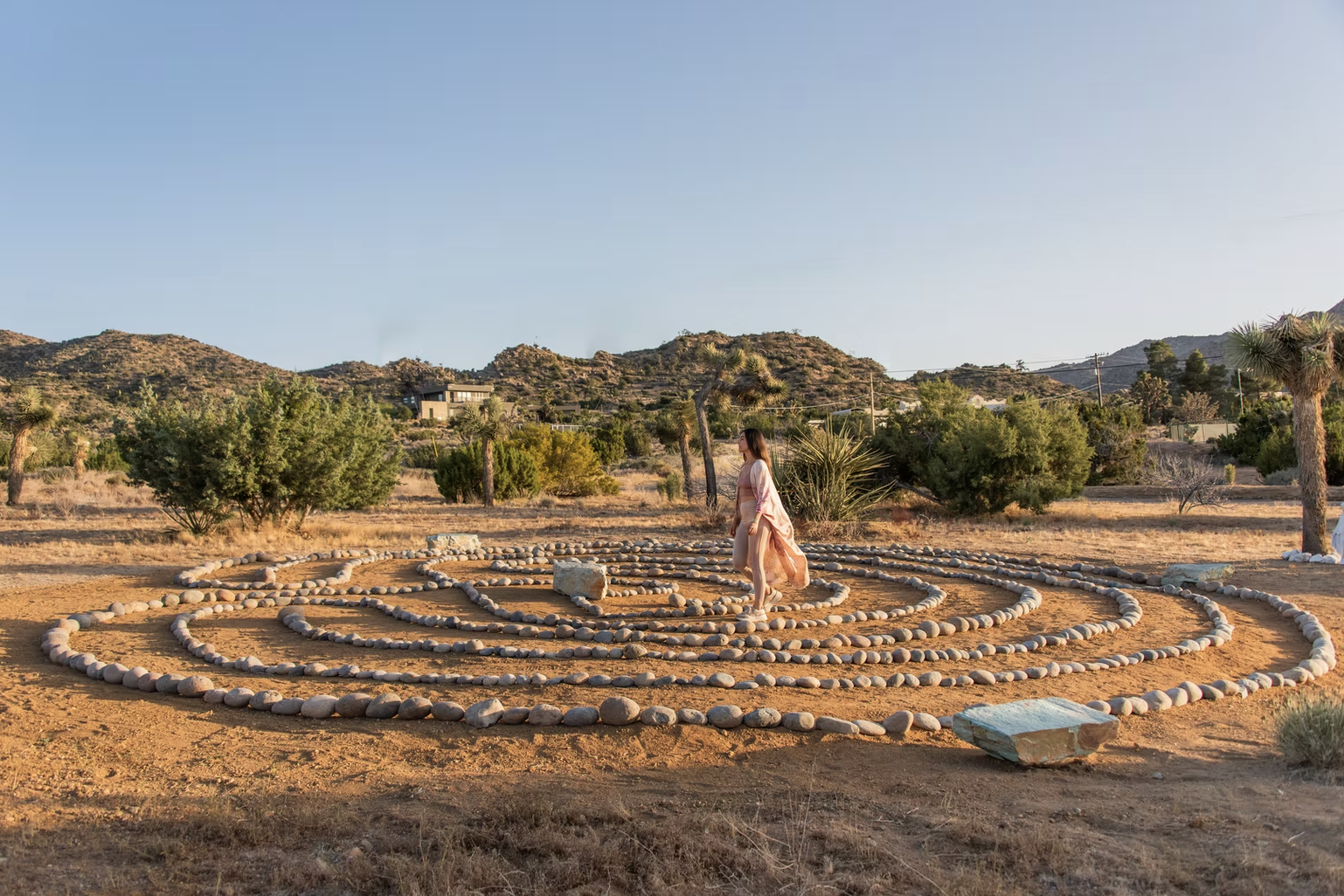 A woman dressed in light clothing walking on a circular stone labyrinth in a desert landscape with mountains, sparse trees, and clear blue sky.