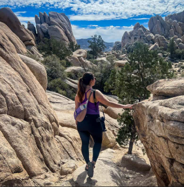 A woman hiking on a rocky trail in a desert-like landscape with large rock formations and sparse trees under a partly cloudy sky.