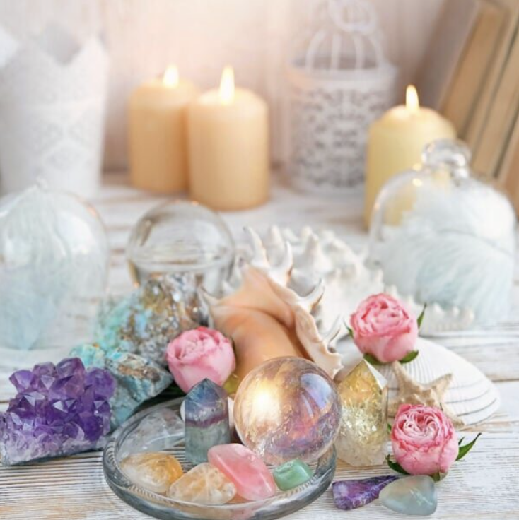 Arrangement of candles, crystals, and pink roses on a white wooden surface, with lit candles in the background.