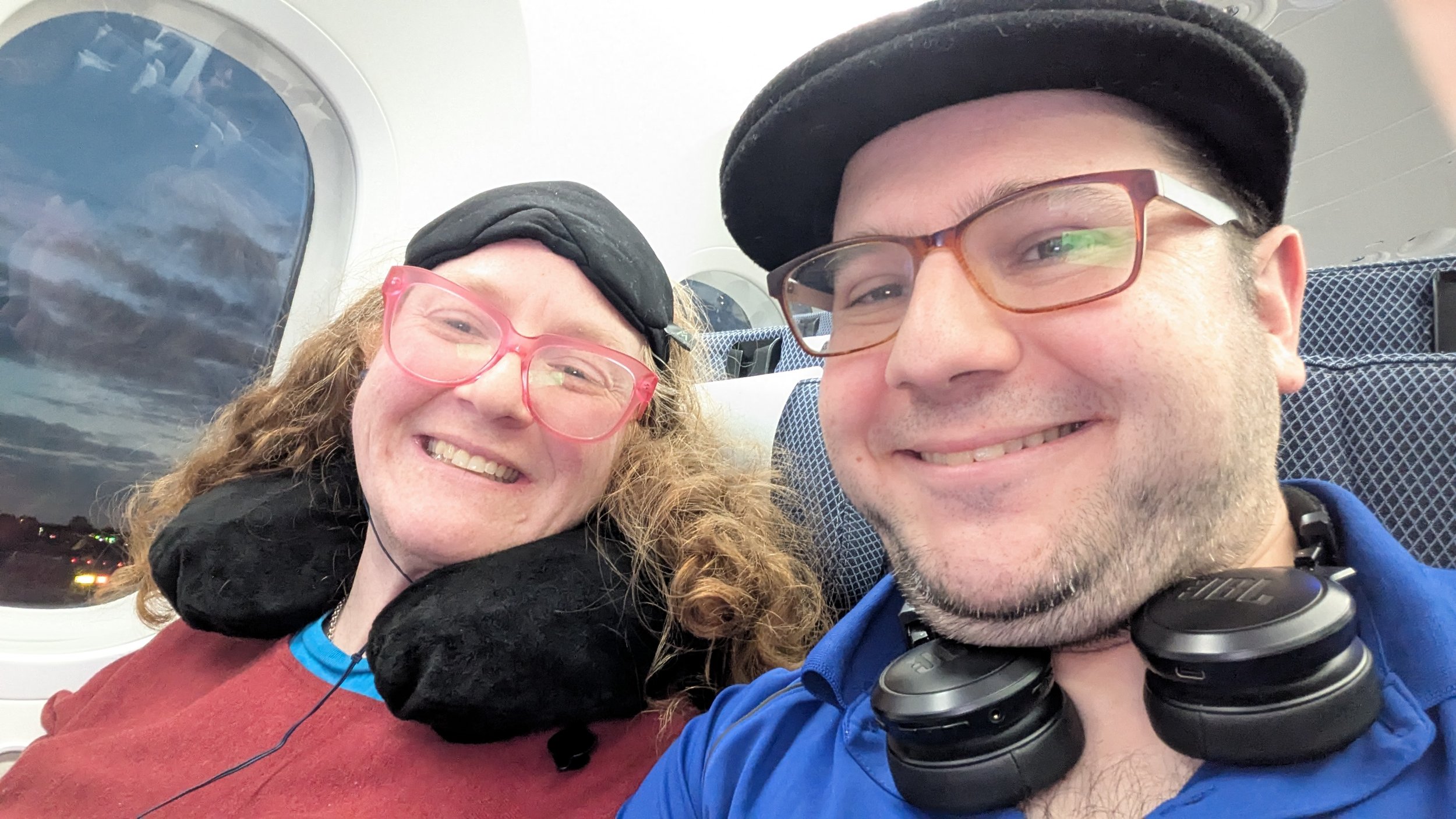 A smiling man and woman taking a selfie inside an airplane, with the sky visible through the window.
