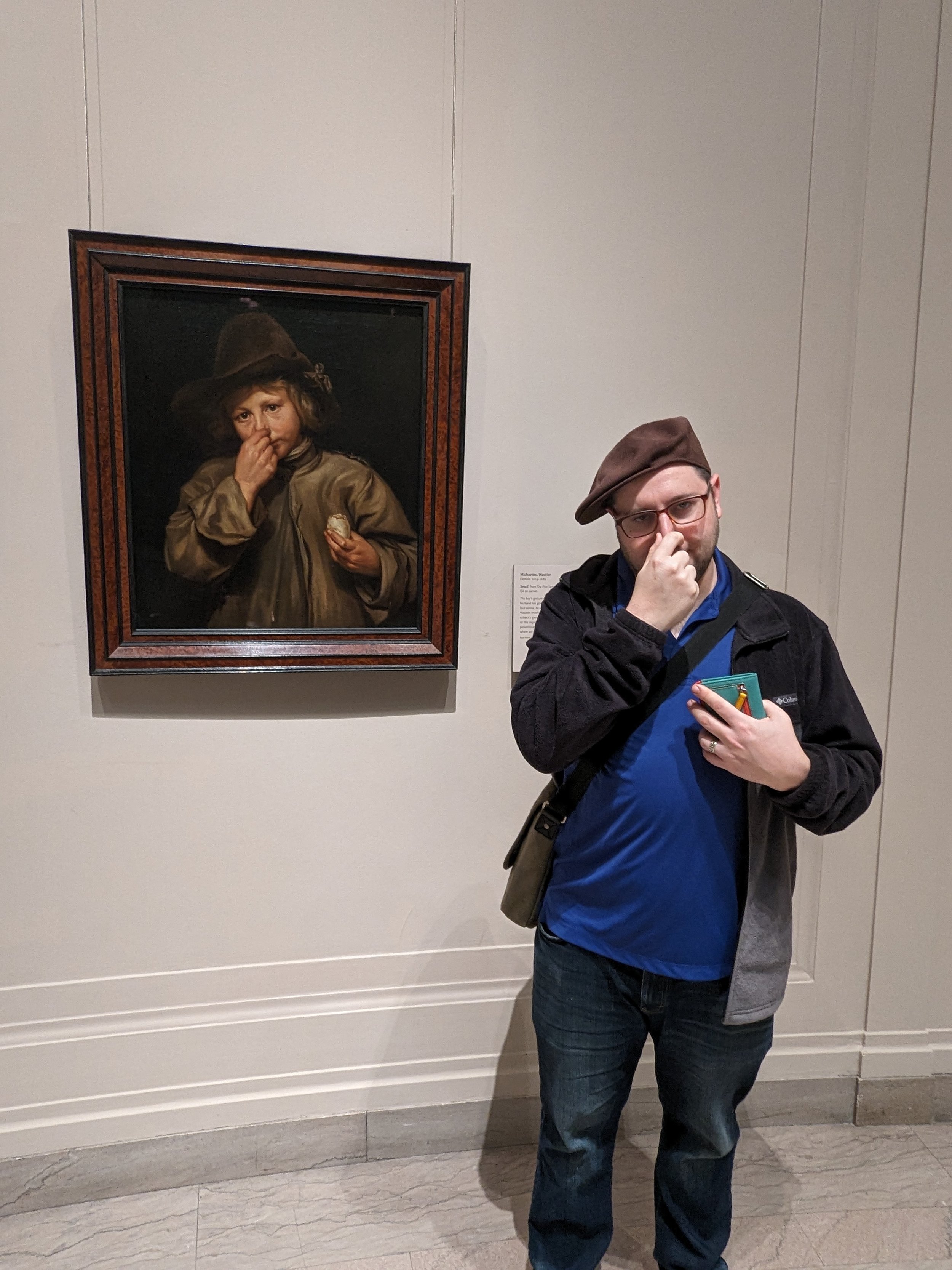 Person standing in an art gallery, touching his nose, with a painting of a young child in historical clothing displayed on the wall behind him.