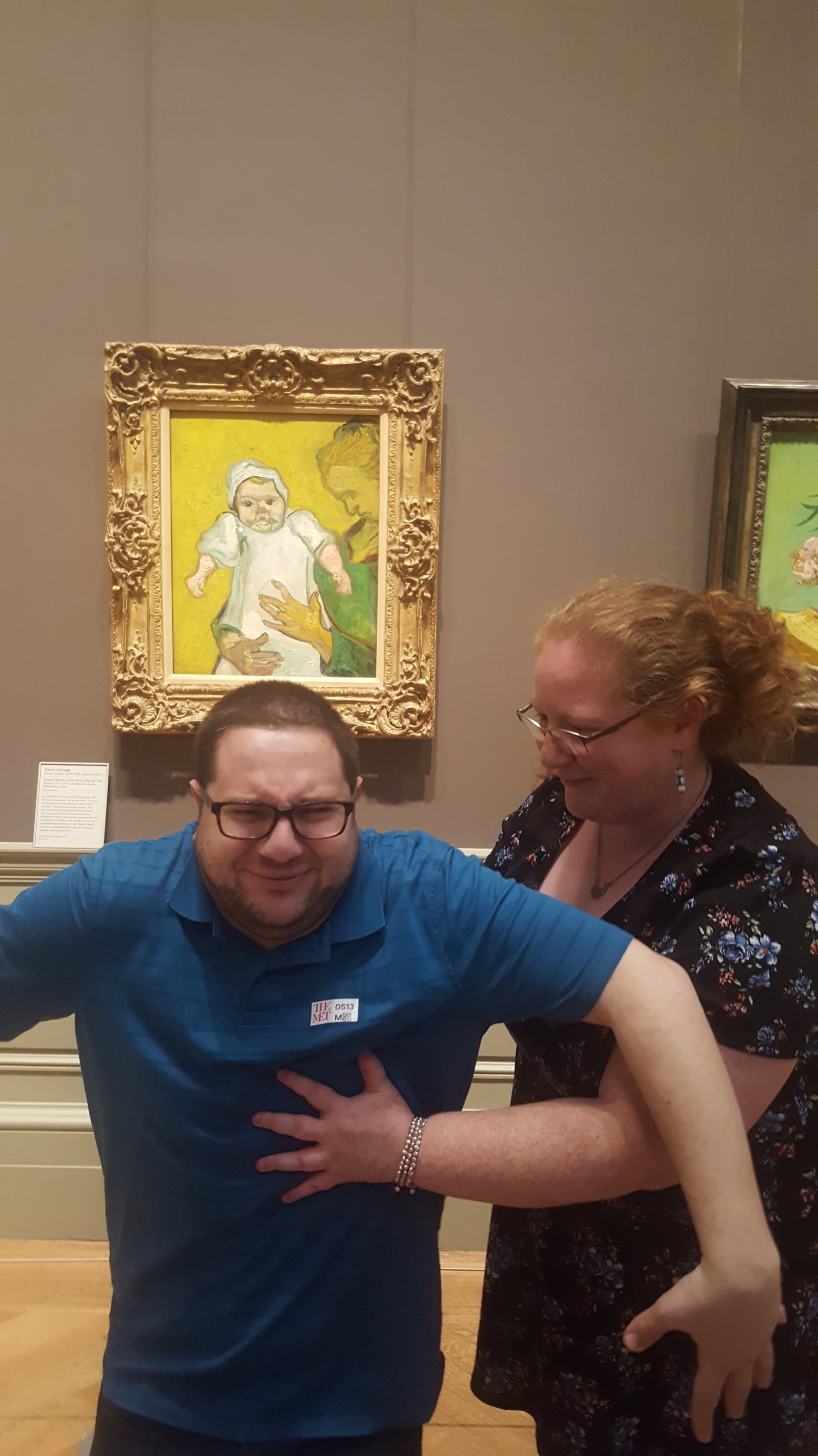 A man appears to be pretending to have a heart attack as a woman mockingly performs CPR in an art museum, in front of a framed painting of a person holding a baby.
