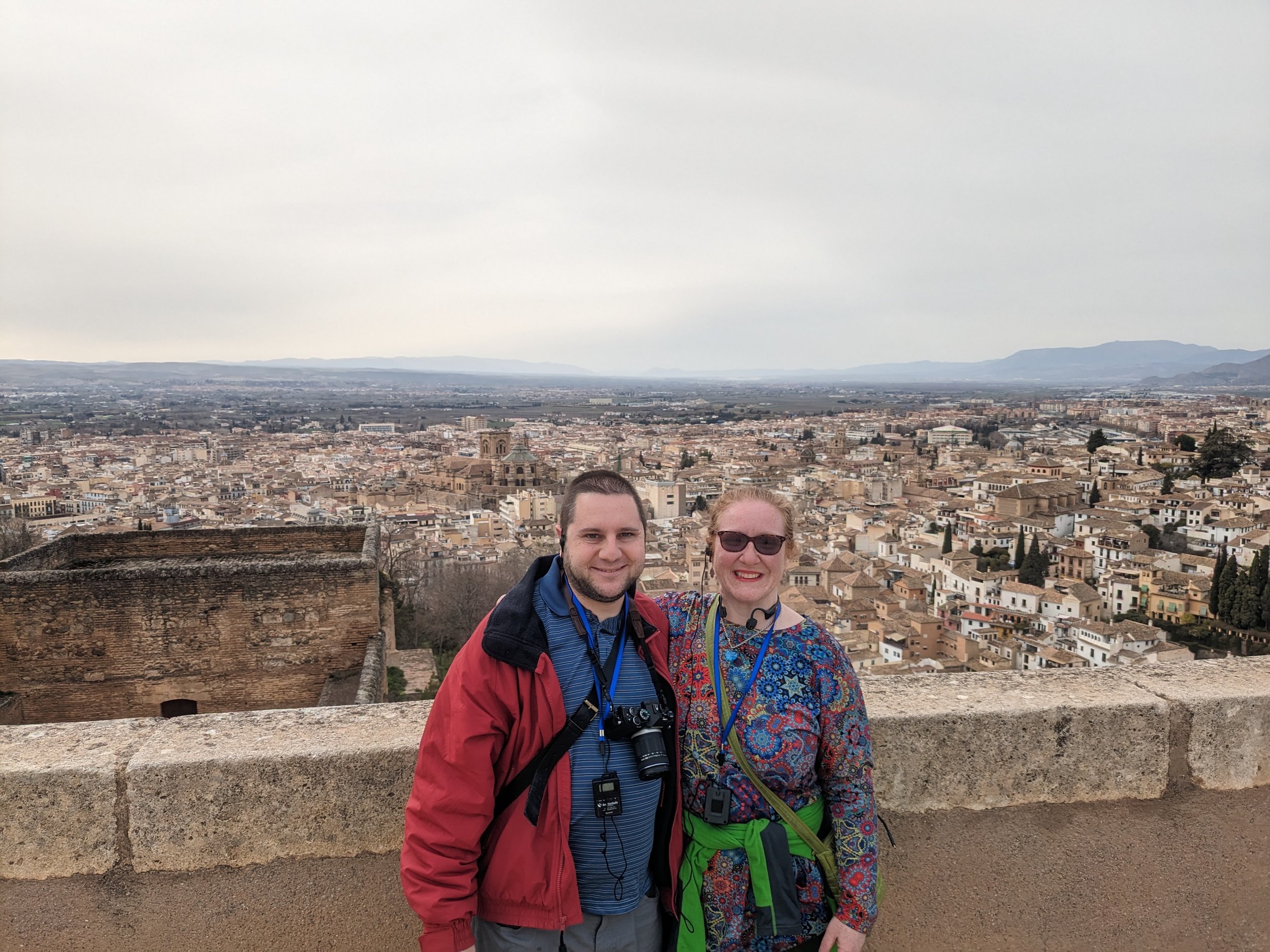 A man and a woman standing on a stone wall overlooking a panoramic view of a historic city with many buildings and distant mountains.