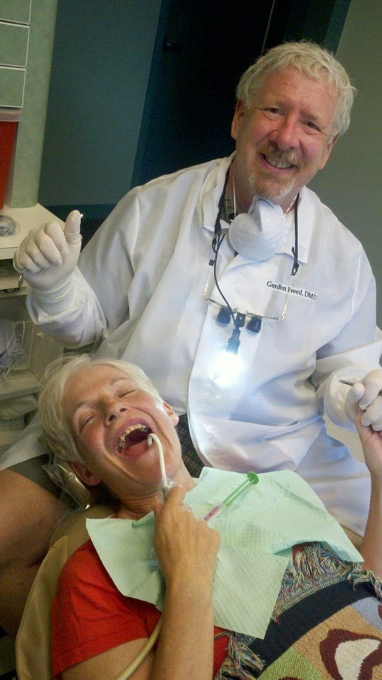A smiling male dentist standing over a woman patient lying in a dental chair, who is laughing with her mouth open, showing dental tools and equipment.