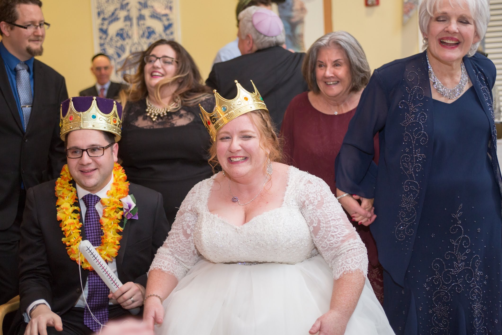 A group of people at a celebration, with a woman in a wedding dress and a man in a suit wearing crowns and leis, indicating a joyful event, likely a wedding with a fun, crowned toast.