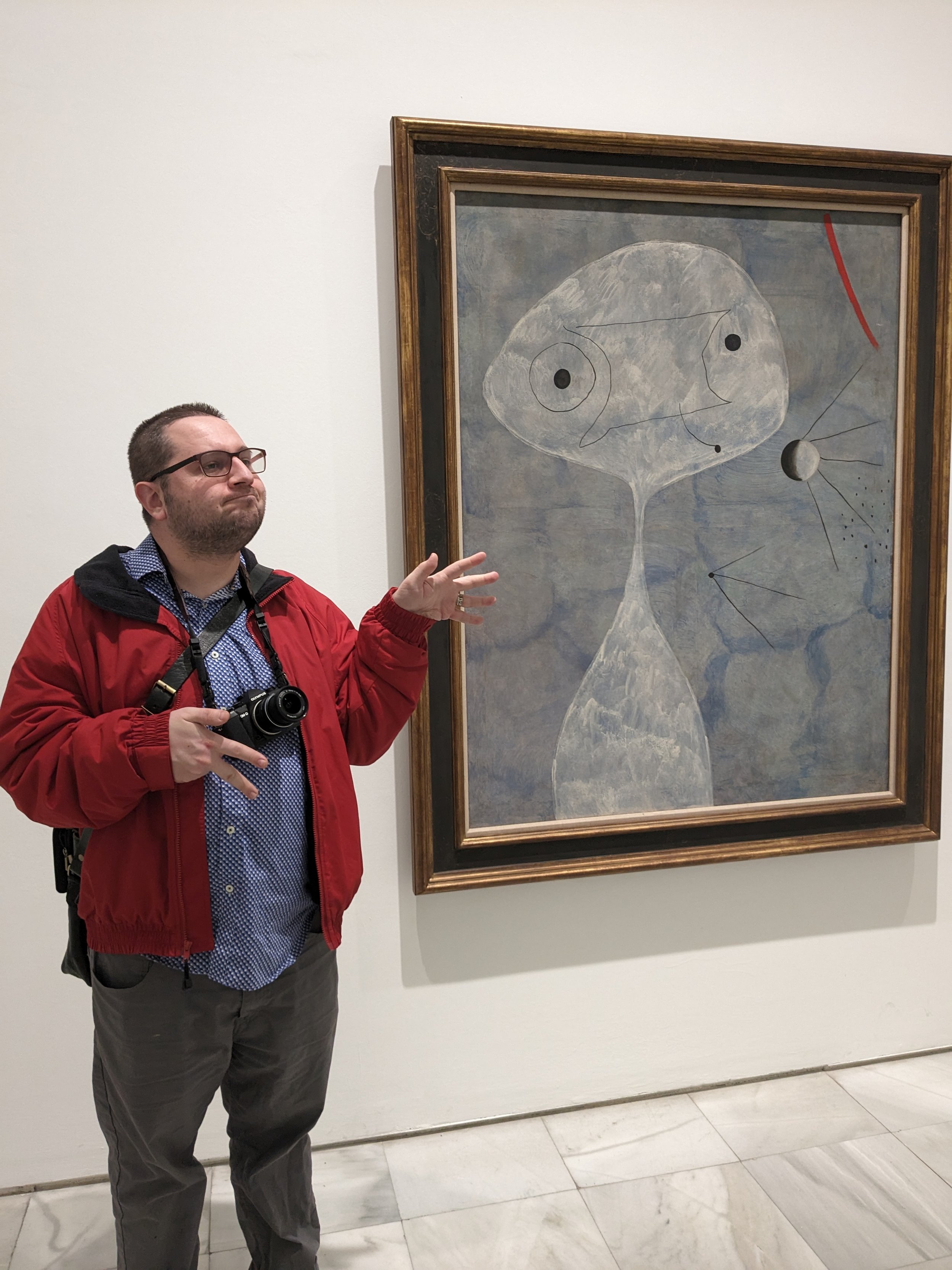 A man with glasses, a beard, wearing a red jacket and carrying a camera, standing beside an abstract painting in a museum or art gallery.