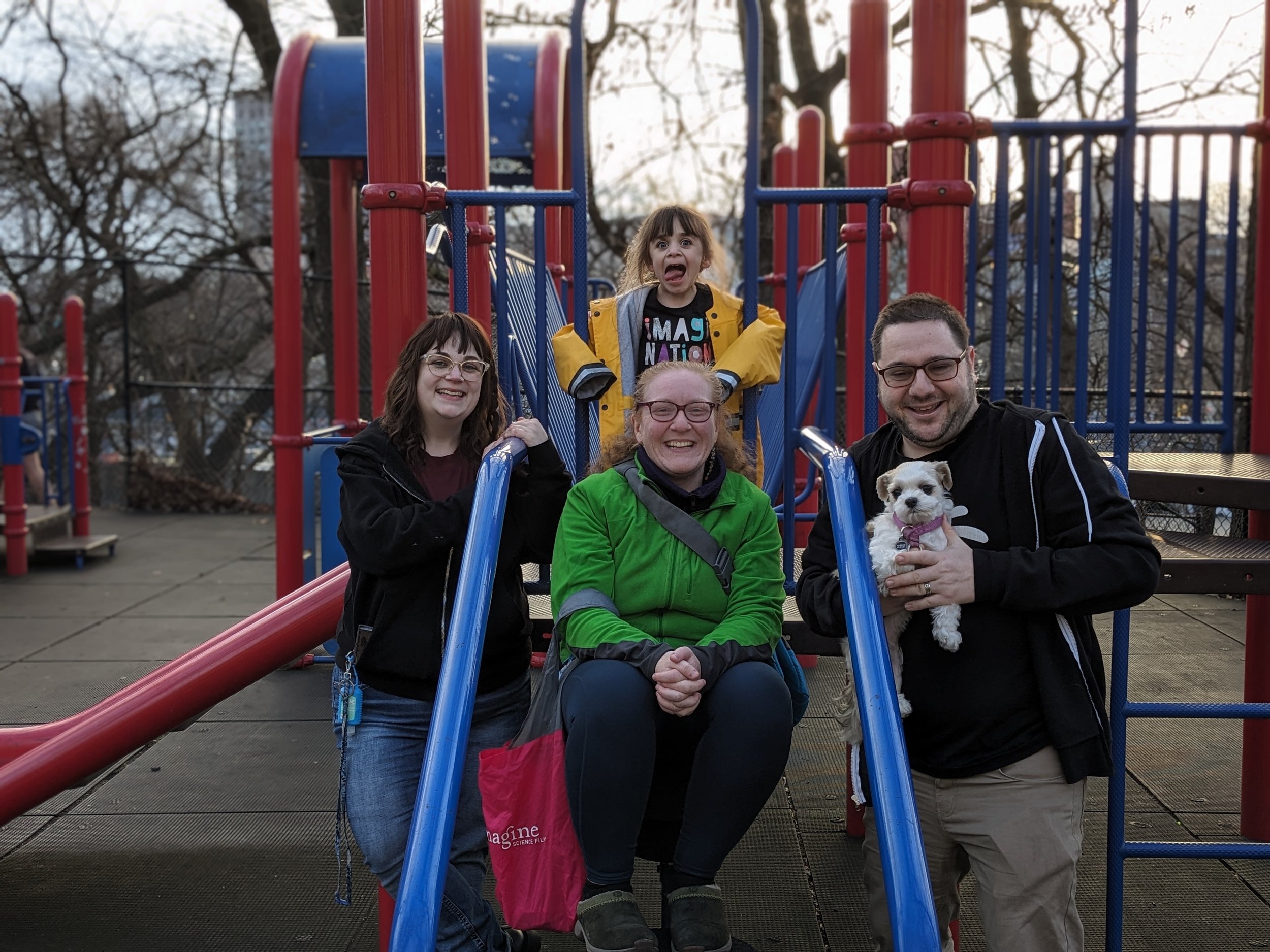 Group of four people and a dog at a playground, with one child on the slide and three adults posing with the dog, all smiling, outdoor setting with trees in the background.