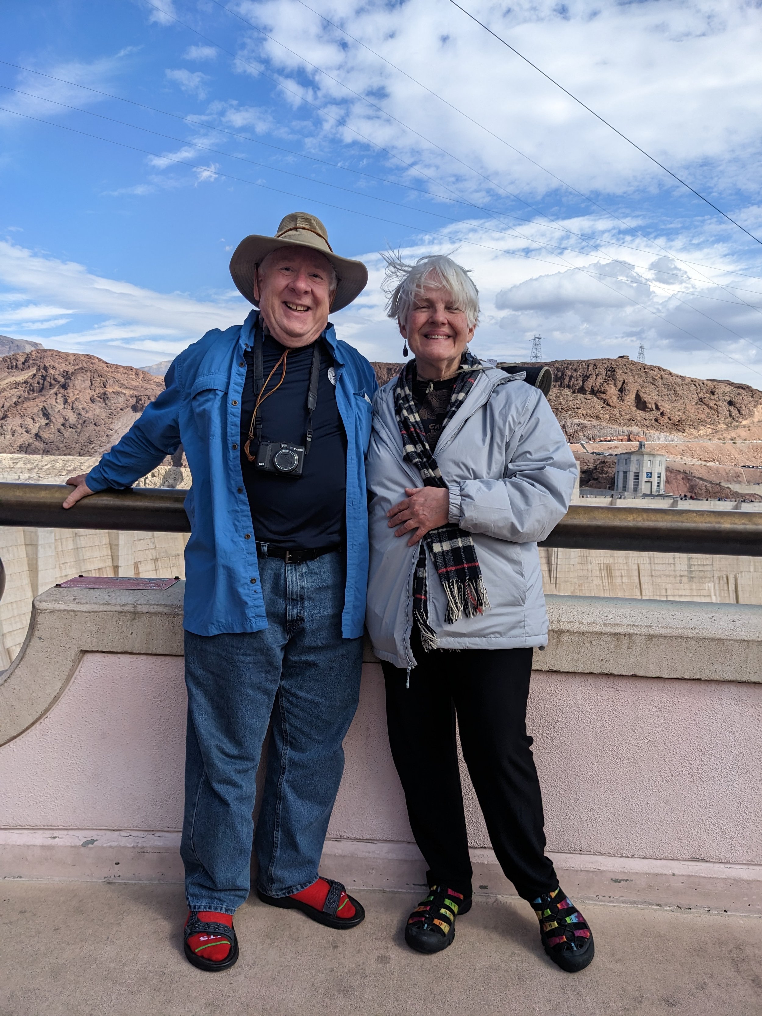 A smiling elderly man and woman standing together outdoors near the Hoover Dam, with rocky mountains and a partly cloudy blue sky in the background.