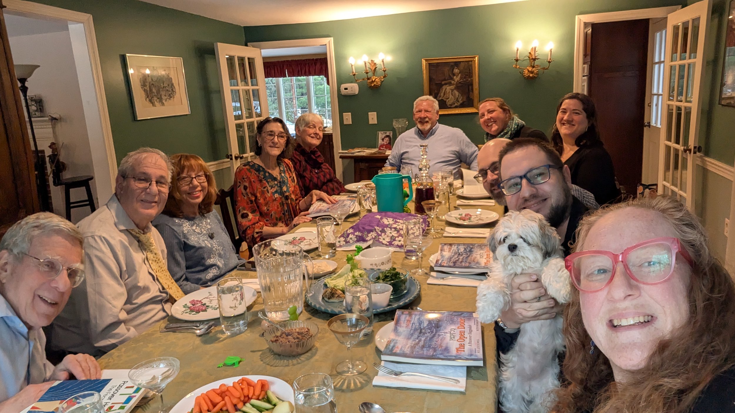 A group of people gathered around a dining table, smiling for a selfie at a dinner party. There is a dog held by a woman with red glasses in the foreground, and the table is set with food, drinks, and books. The background shows green walls, wall-mou