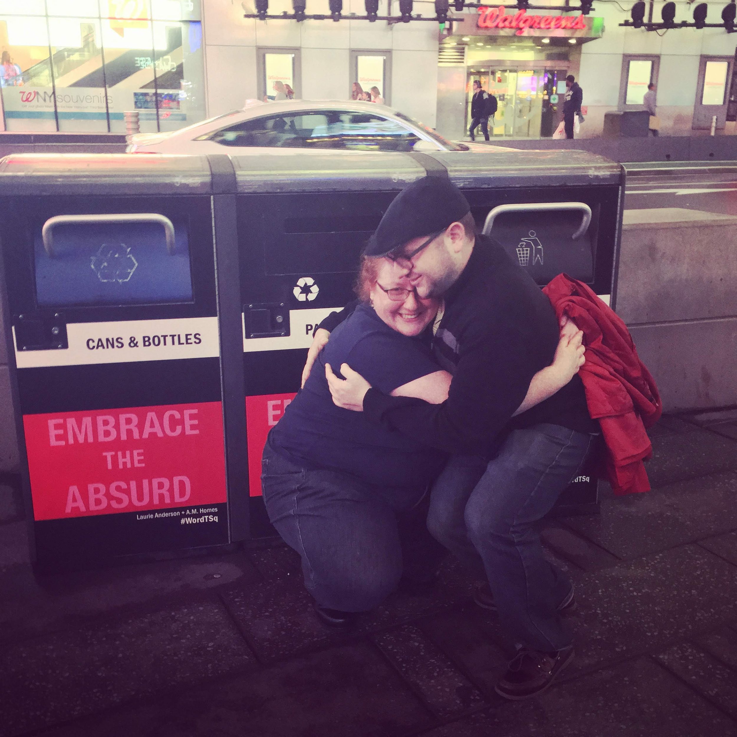 Two people hugging and laughing on the sidewalk at night, next to a recycling bin that has a sign saying "Embrace the Absurd." They are outside a Walgreens store with people walking by in the background.
