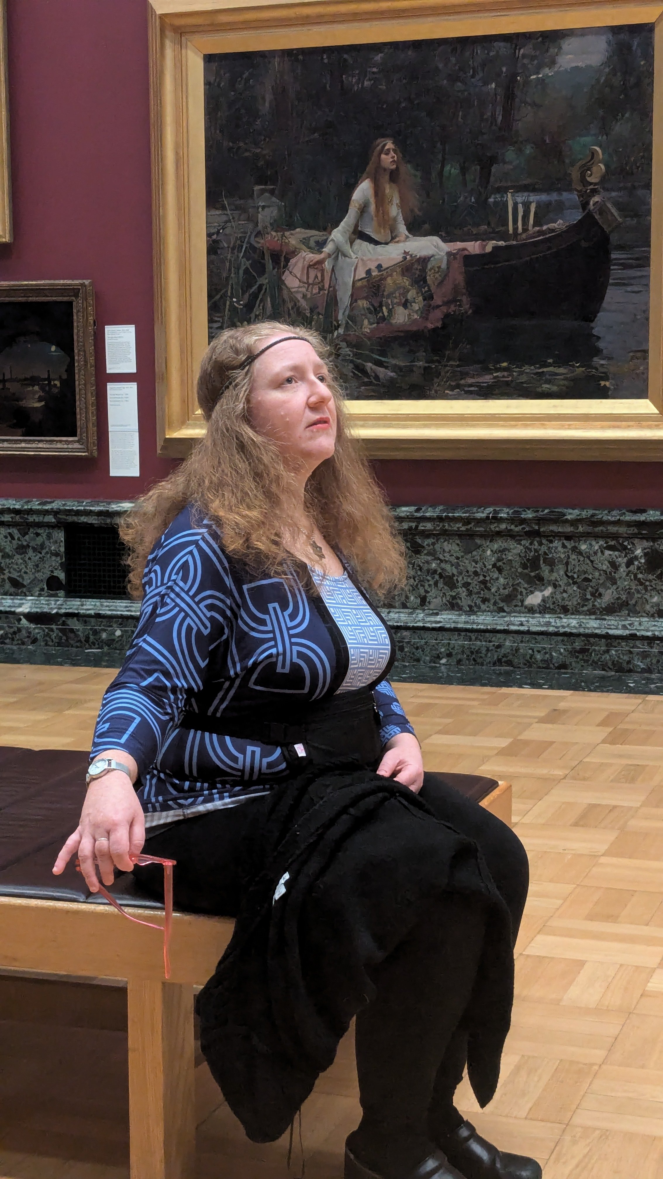 A woman with long, wavy hair wearing a blue patterned jacket and black pants sitting on a bench in an art gallery, with a large painting of a woman on a boat behind her.