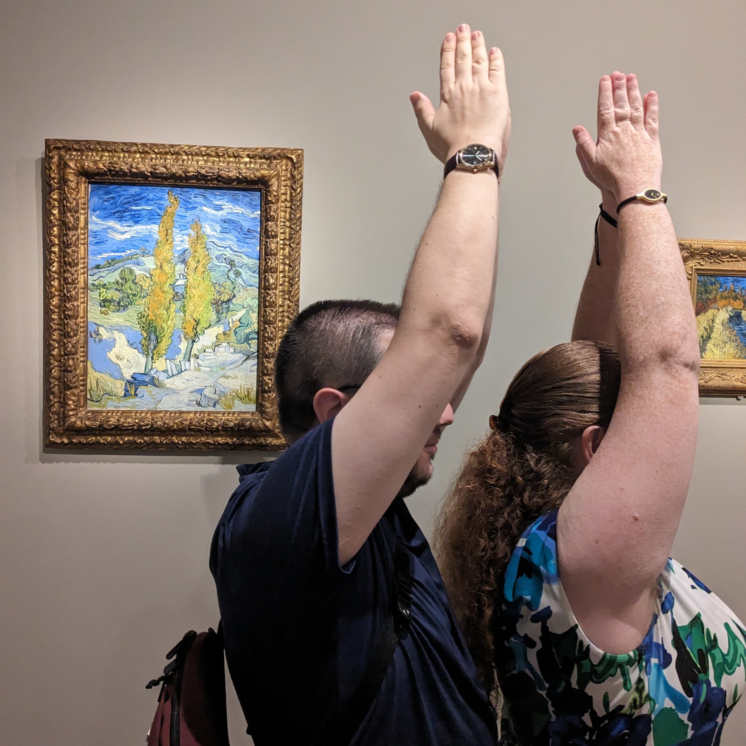 Two people with arms raised, standing side by side in an art gallery, with a Vincent van Gogh painting of cypress trees and a landscape hanging behind them.