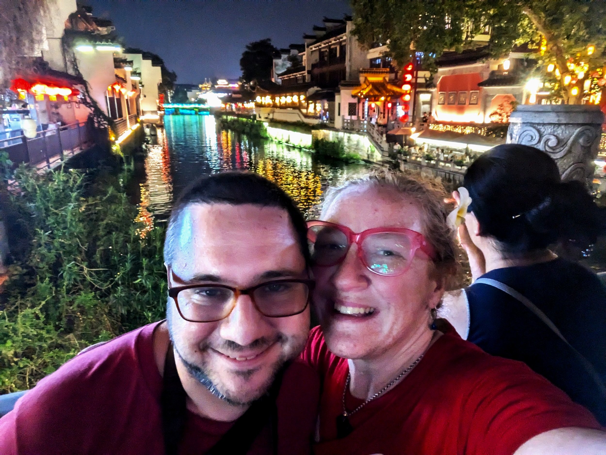 A smiling couple taking a selfie outdoors at night with a lit-up canal and traditional buildings in the background.