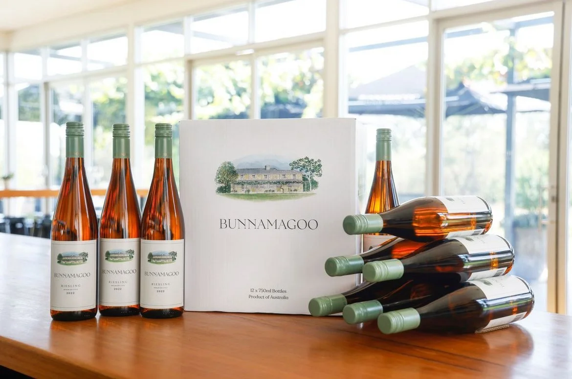 Six bottles of Bunnamagoo Riesling wine and a box of 12 bottles on a wooden table inside a bright room with large windows.