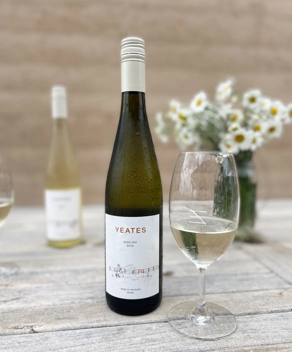A bottle of Yeates Riesling 2022 wine with a glass of white wine on a wooden table, with a vase of white daisy flowers in the background.