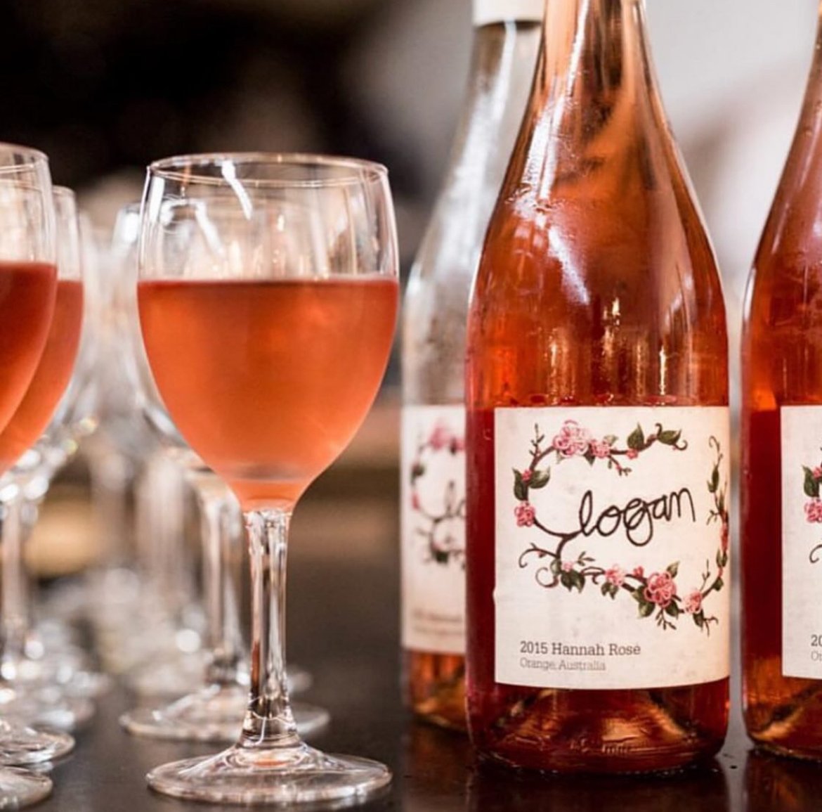 Several glasses of rosé wine and bottles with custom floral labels, with one bottle labeled "Logan" and "2015 Hannah Rosé" from Orange, Australia.