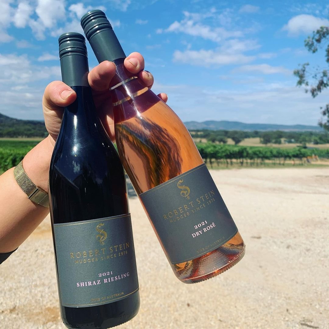 Hand holding two bottles of wine, one Shiraz Riesling and one Dry Rosé, against a rural landscape with fields, trees, and hills under a blue sky with some clouds.