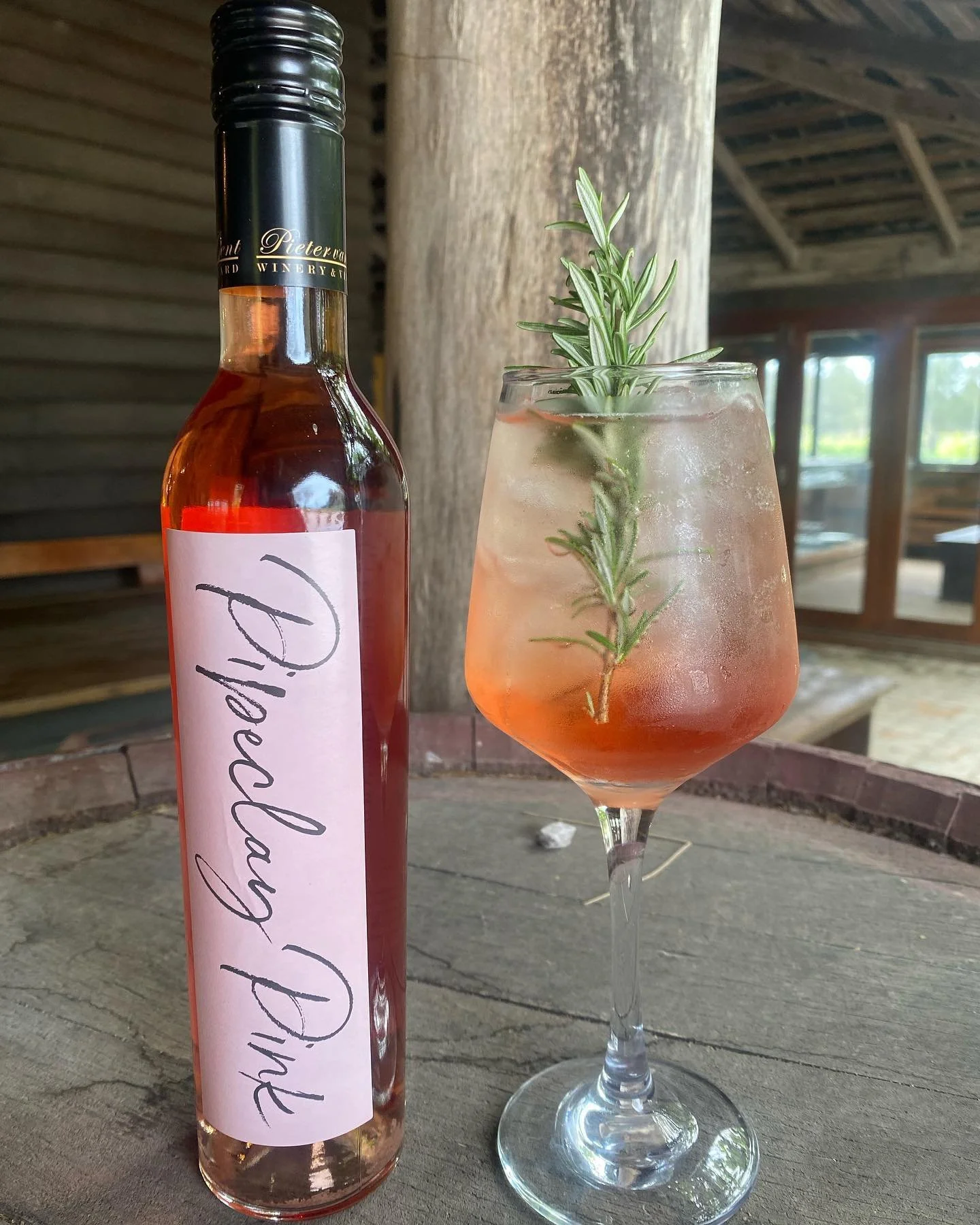 A bottle labeled 'Pellowny Dye' and a cocktail glass with a pinkish drink, ice, and a sprig of rosemary garnish, set on a rustic wooden surface in a barn-like setting with windows in the background.