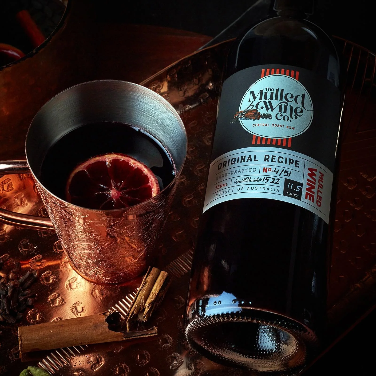 A bottle of mulled wine with a label reading 'The Mulled Wine Co.', next to a glass of dark red mulled wine with a slice of blood orange inside, on a reflective copper surface with some spices and cinnamon sticks.