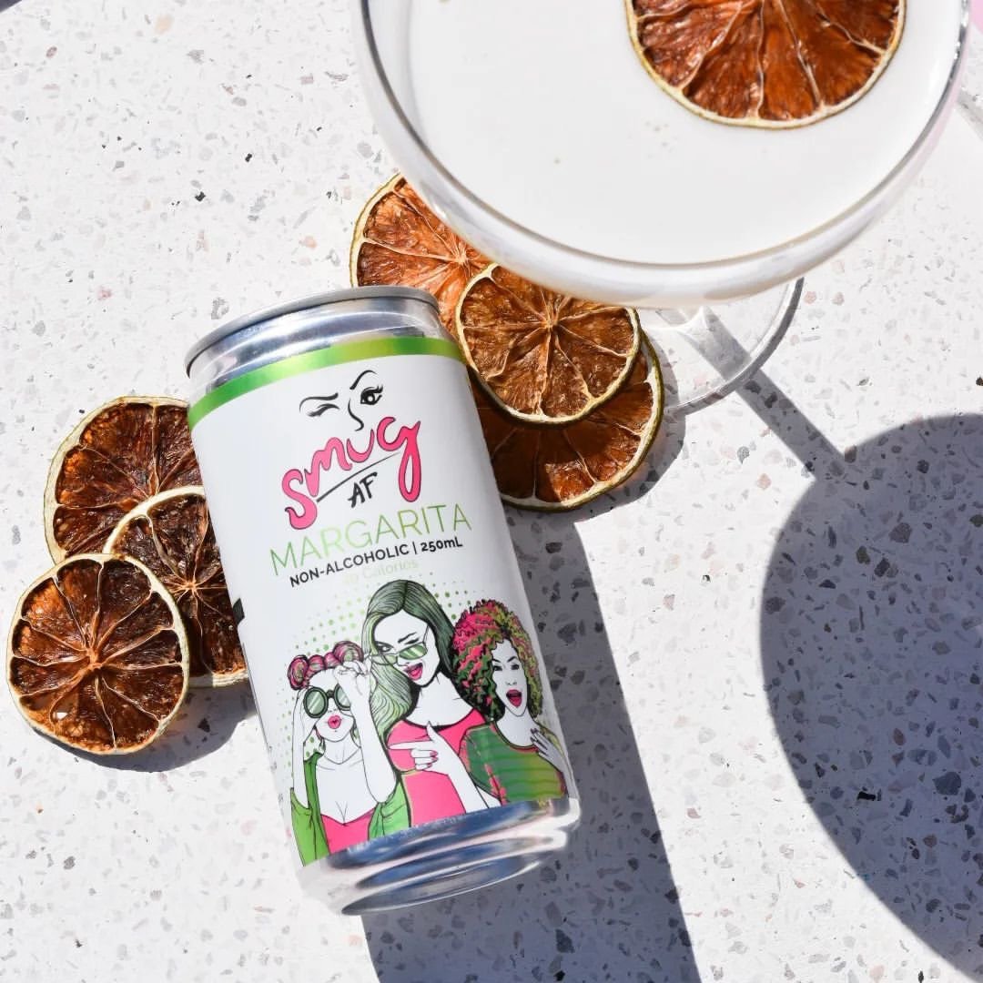 Can of Smug AF Margarita non-alcoholic beverage beside dried citrus slices and a cocktail glass on a white speckled surface.