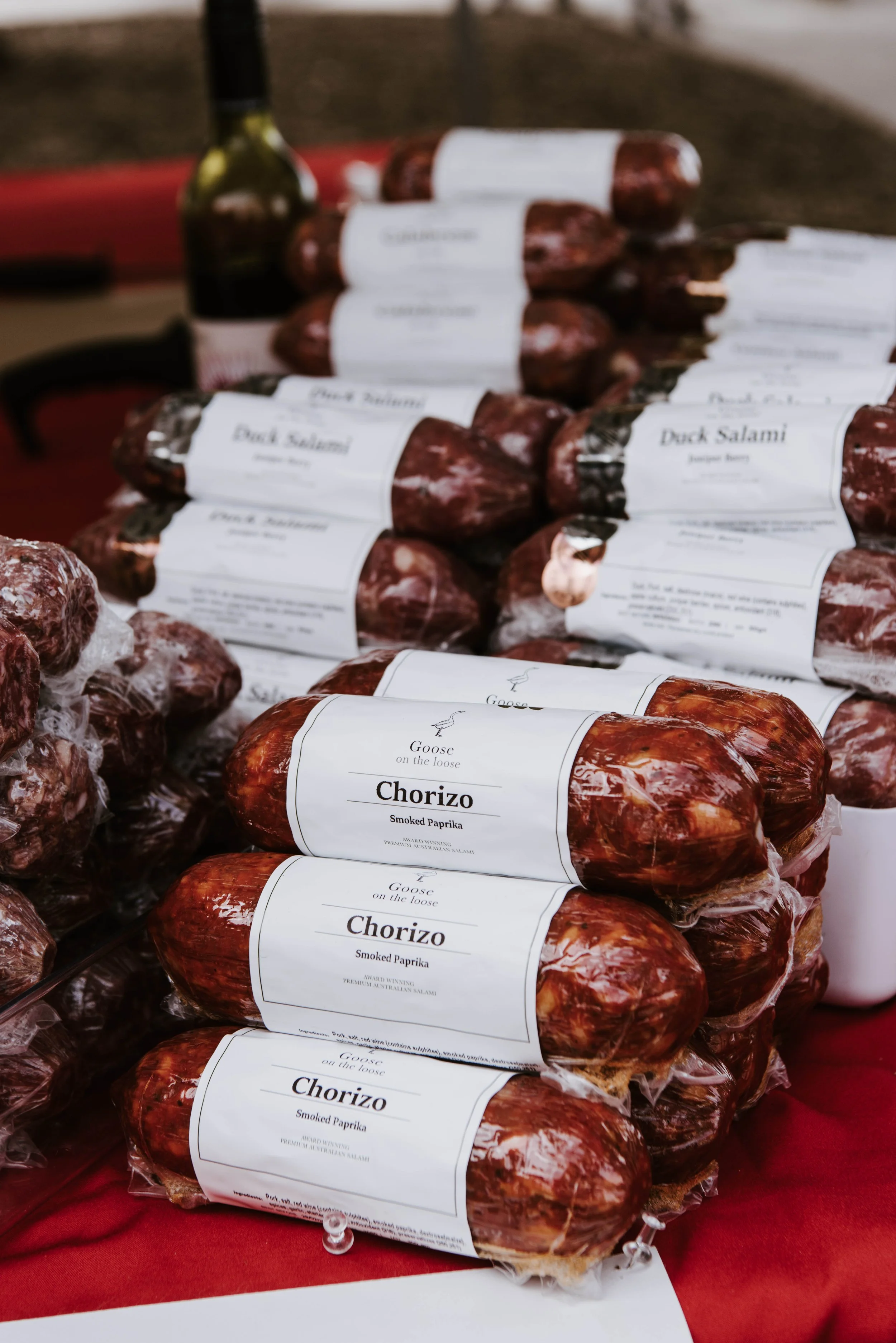 Stacked packages of smoked chorizo sausage labeled "Goose on the loose" at an outdoor market stall with other smoked meats in the background.