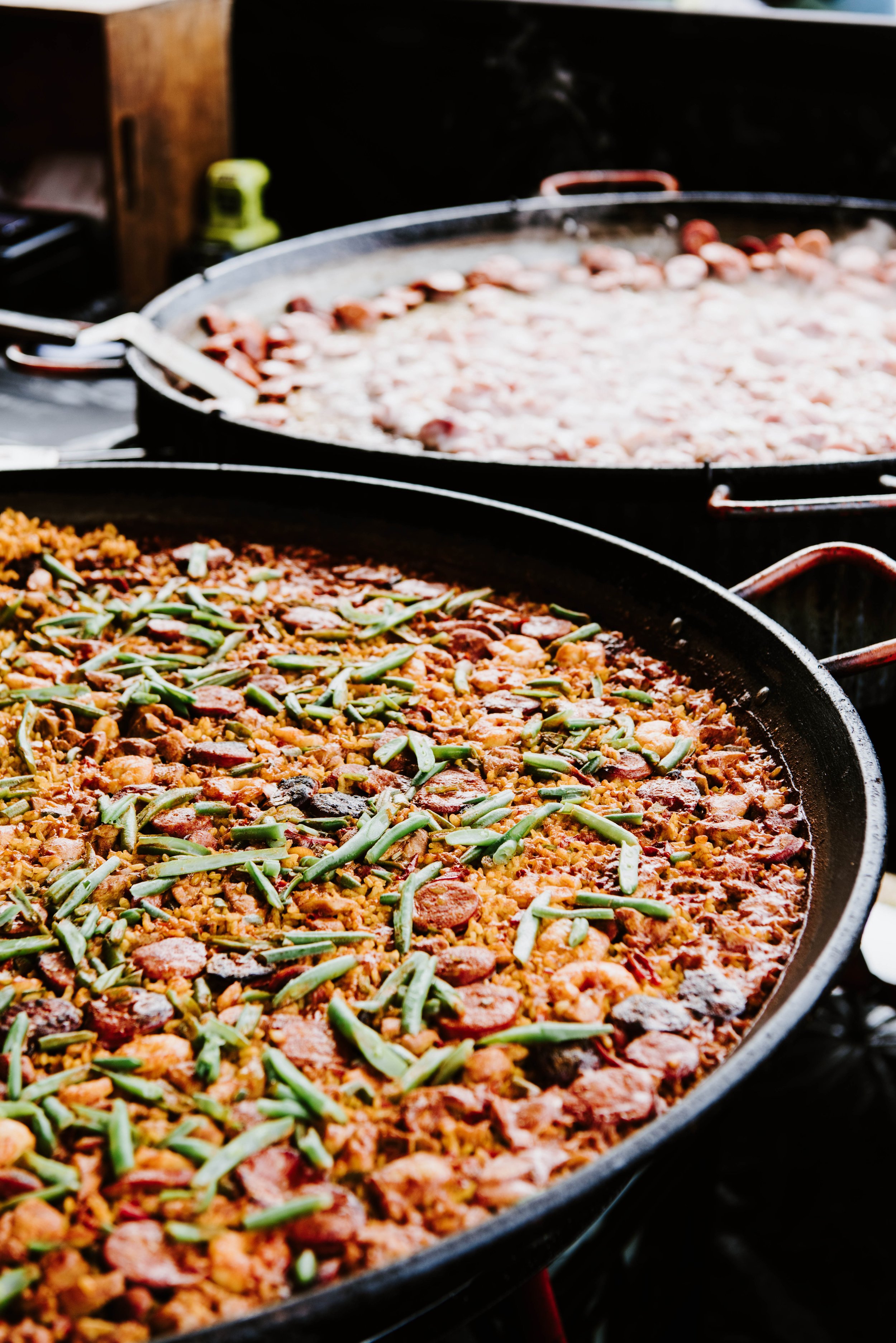 Large outdoor pans of cooked paella with rice, green beans, slices of sausage, and other ingredients.