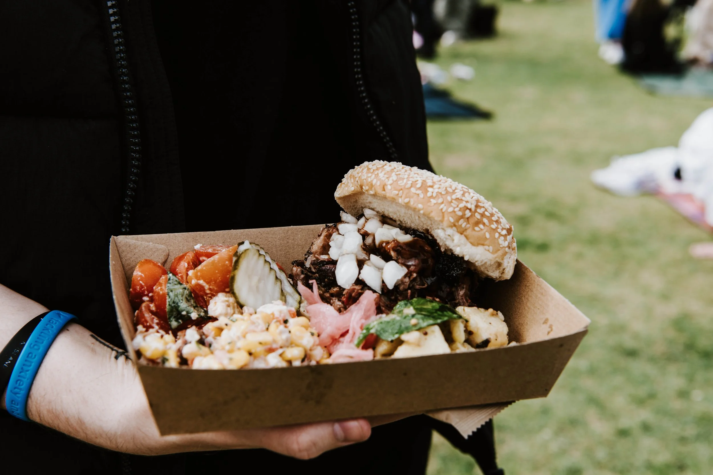Person holding a cardboard tray with a burger topped with chopped nuts, grilled vegetables, a pasta salad, and sliced pickles.