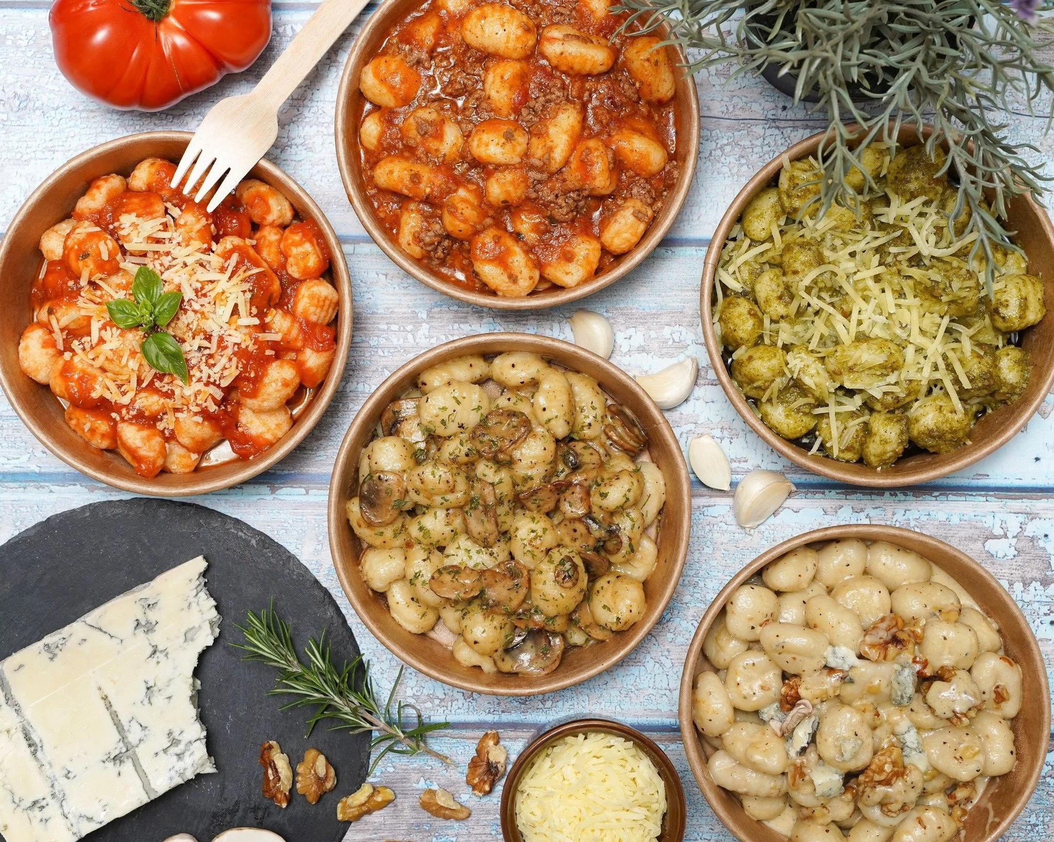 Assorted bowls of gnocchi with different sauces, cheese, and herbs on a rustic table with garlic, walnuts, and blue cheese.