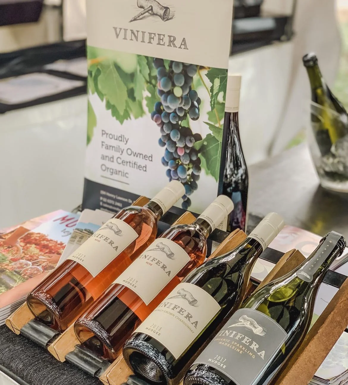 Display of bottles of Vinifera wine, including rosé, Chardonnay, and sparkling wine, in front of a poster with grapes and a vineyard scene, promoting organic, family-owned wine from Mudgee.