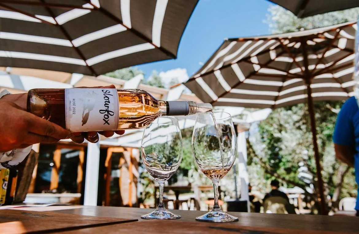 A person pours rosé wine into two empty wine glasses on a wooden table outdoors under a striped umbrella on a sunny day.