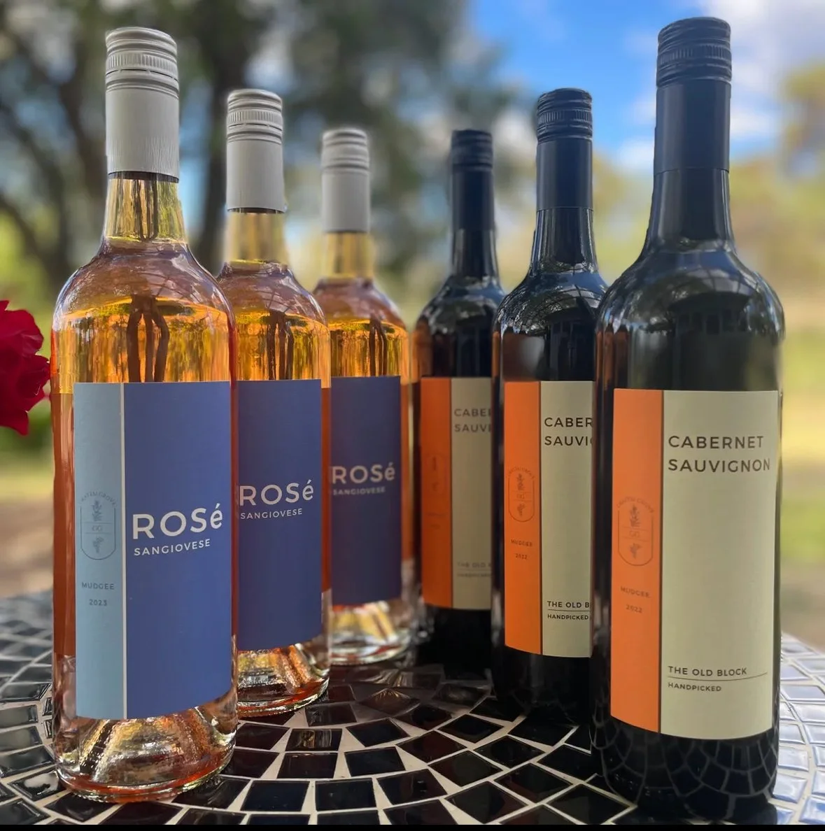 Six bottles of wine, three rosé and three cabernet sauvignon, arranged on a mosaic table outdoors with trees in the background.