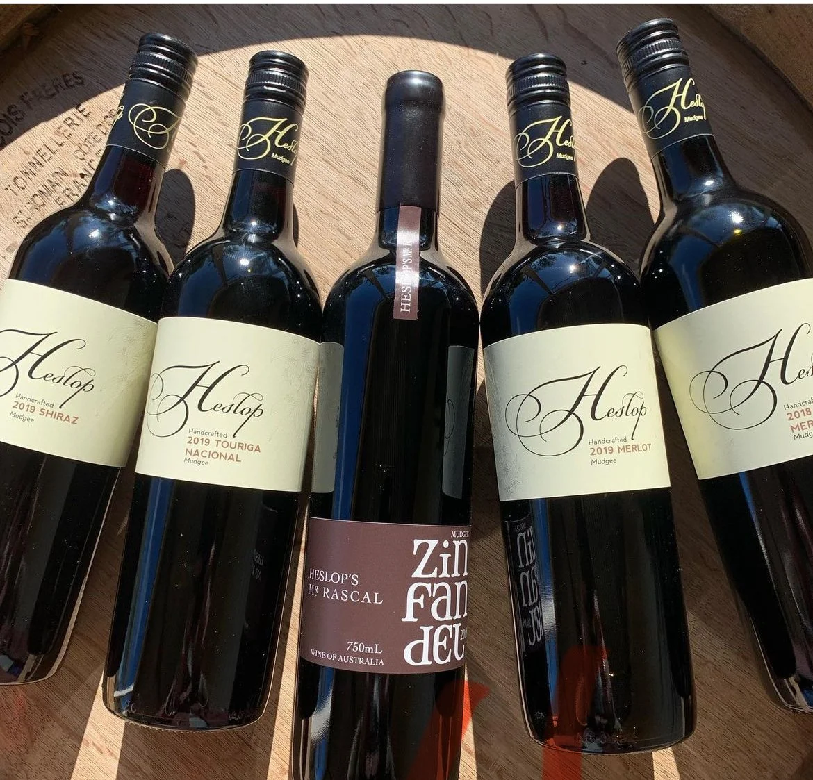 Six bottles of red wine from HESLOP winery, including Shiraz, Touriga Nacional, Merlot, and Mr Rascal Zin Fan pad, arranged on a wooden surface.