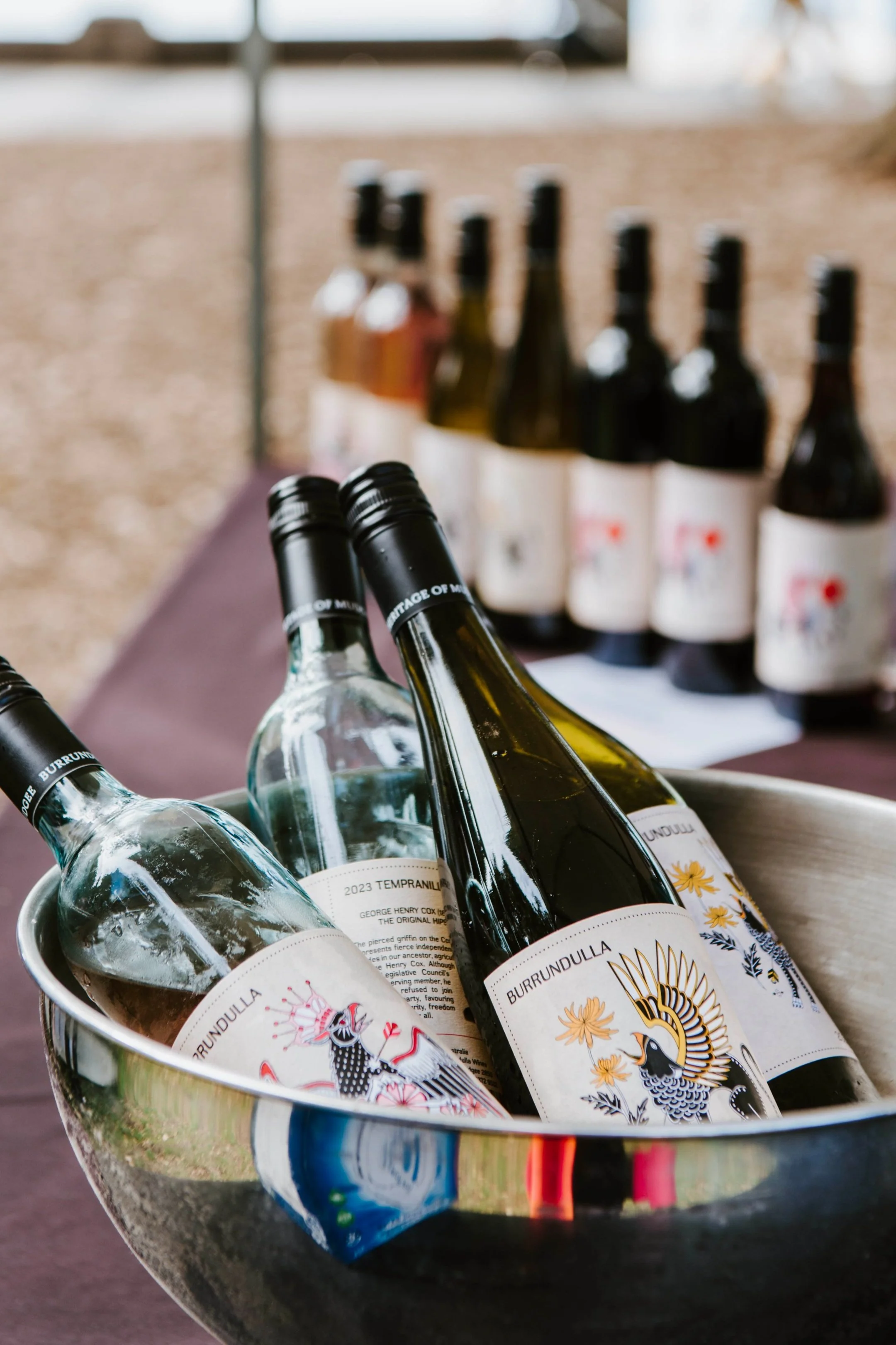 Bottles of wine in a metal bucket at an outdoor event with more bottles in the background on a table.
