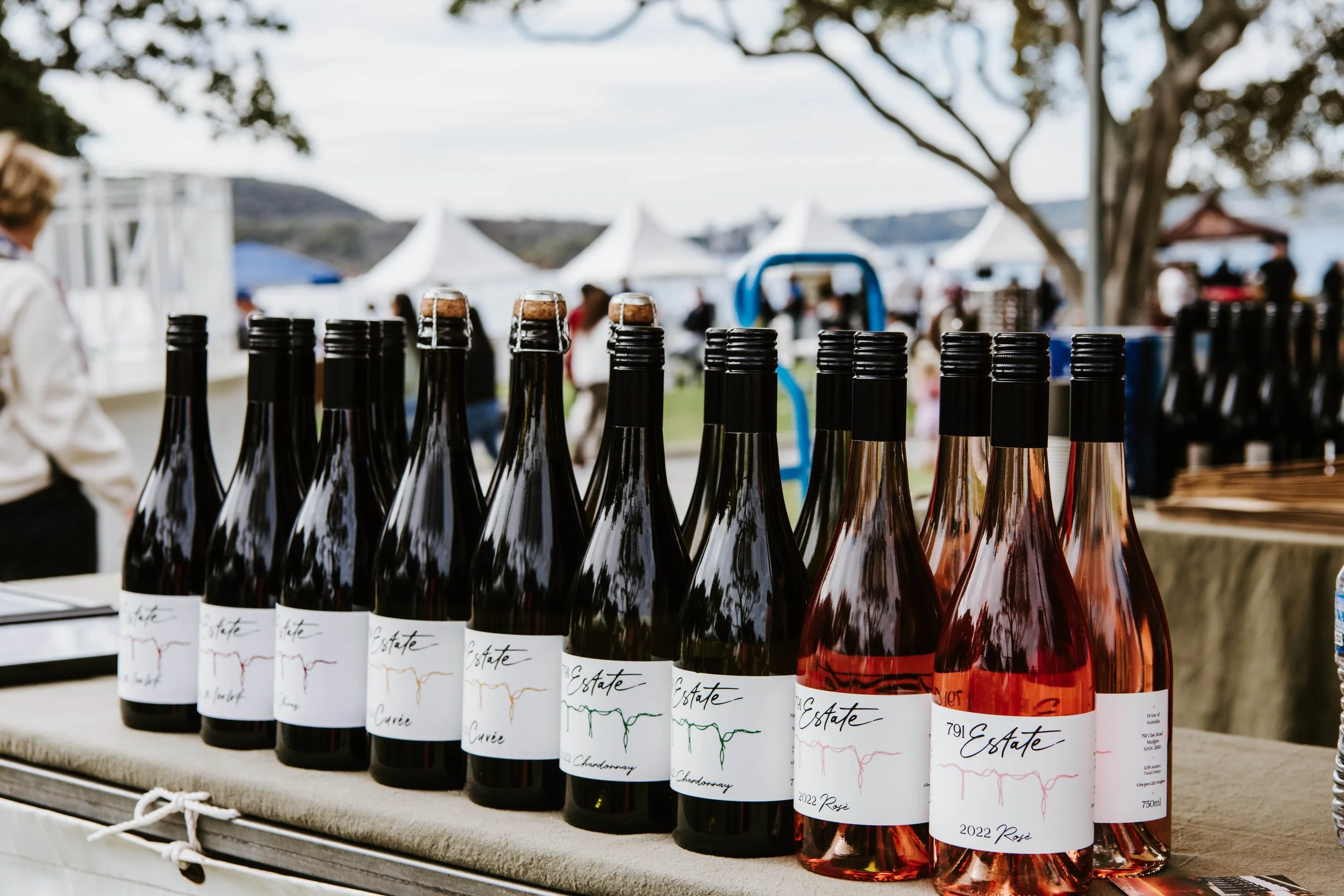 Bottles of wine on a table at an outdoor festival with white tents and people in the background.