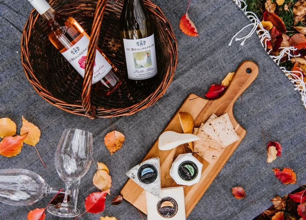 A picnic setup featuring a woven basket with two bottles of wine, a cheeseboard with assorted cheeses and crackers, empty wine glasses, and autumn leaves on a gray woven blanket.