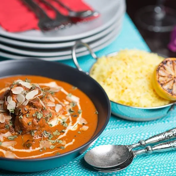 A plate of Indian butter chicken curry garnished with cream, herbs, and almond slices, served with a side of yellow rice and a grilled lemon half.