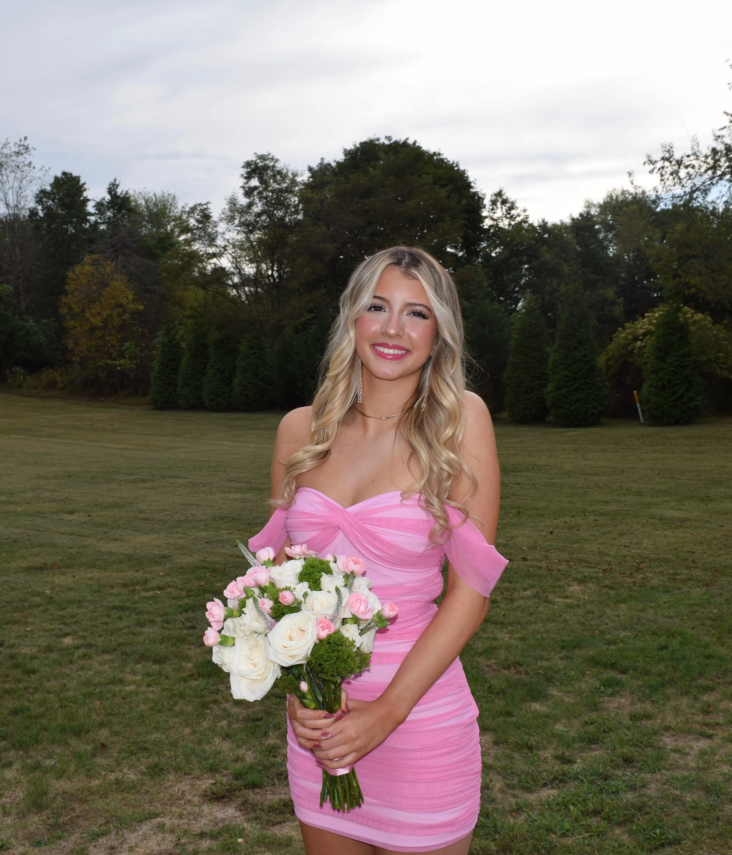 A young woman in a pink formal dress holding a bouquet of flowers, standing outdoors on grass with trees and cloudy sky in the background.