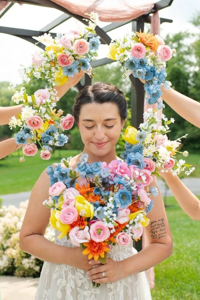 A woman in a wedding dress with her eyes closed, holding a bouquet of colorful flowers, surrounded by a floral wreath being arranged around her head, outdoors with greenery in the background.