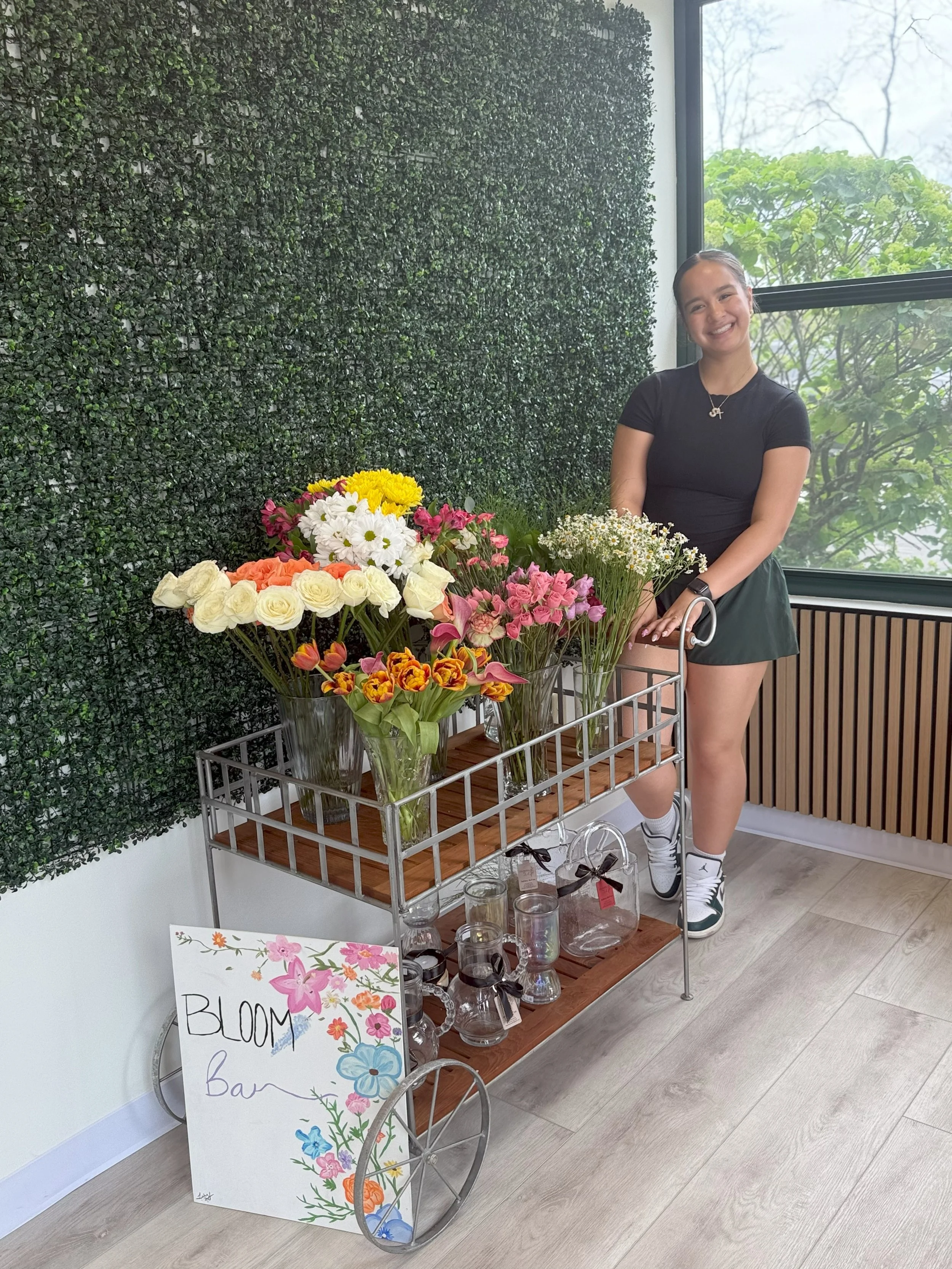 A young woman standing beside a floral cart with a variety of colorful flowers inside, next to a sign reading 'BLOOM Bar.' She is smiling, dressed in a black t-shirt, green shorts, and sneakers, with a green leafy wall and window in the background.