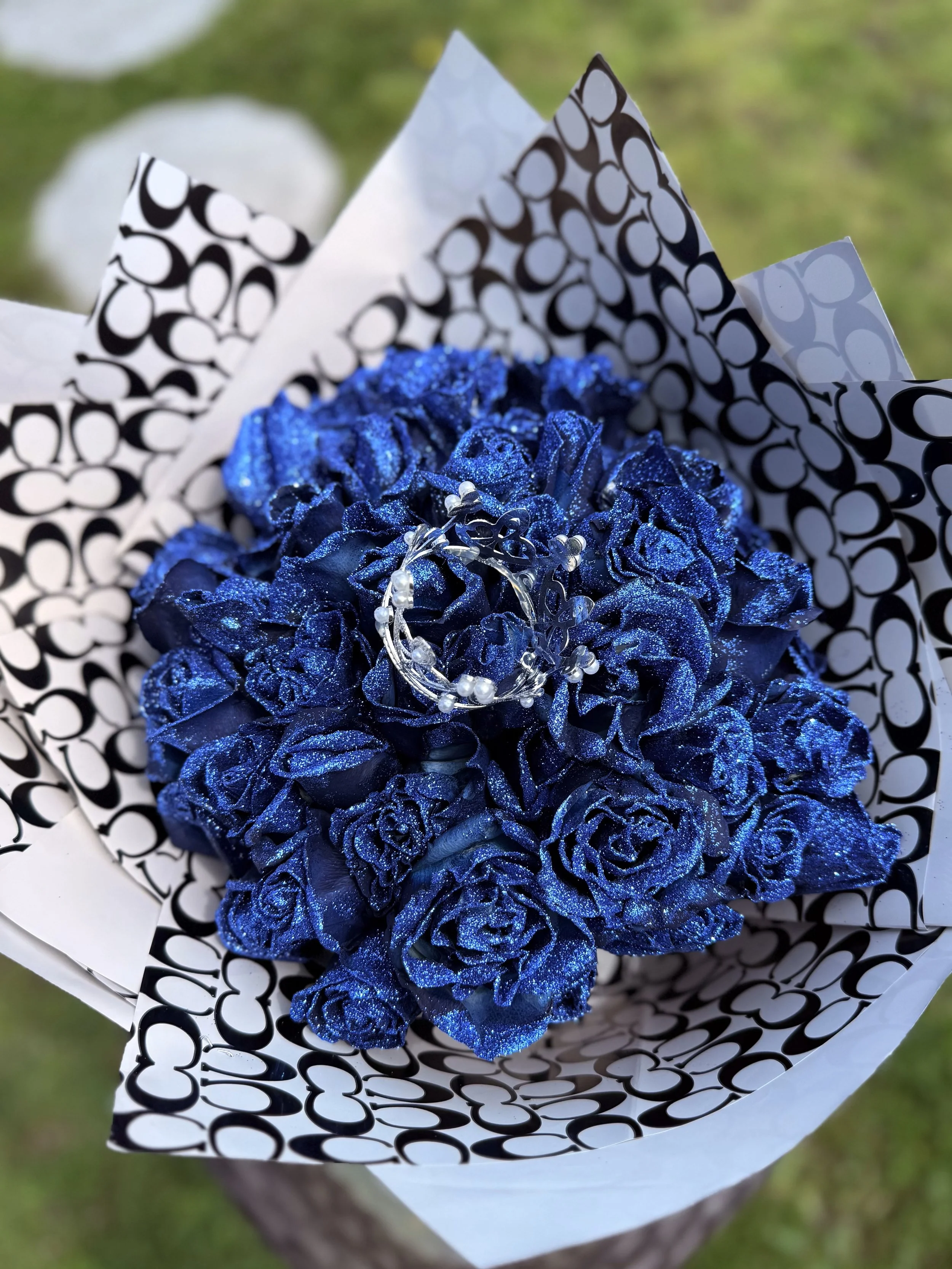 A bouquet of glittery blue roses wrapped in black and white patterned paper, with a silver bracelet placed on top of the roses.