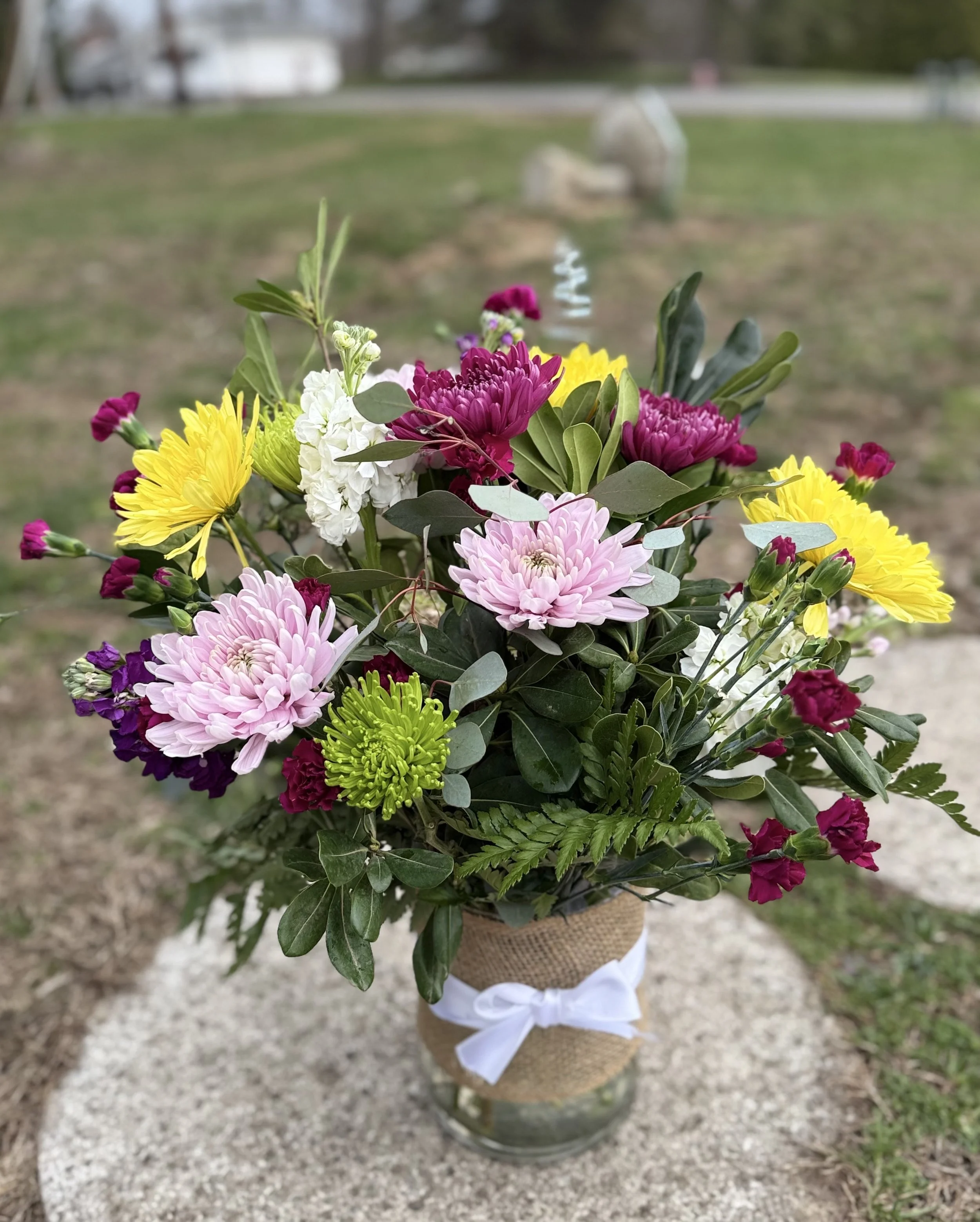 A colorful bouquet of flowers in a vase wrapped with burlap and a white ribbon placed outdoors on a stone surface. The bouquet includes pink, yellow, green, and purple flowers with green foliage.