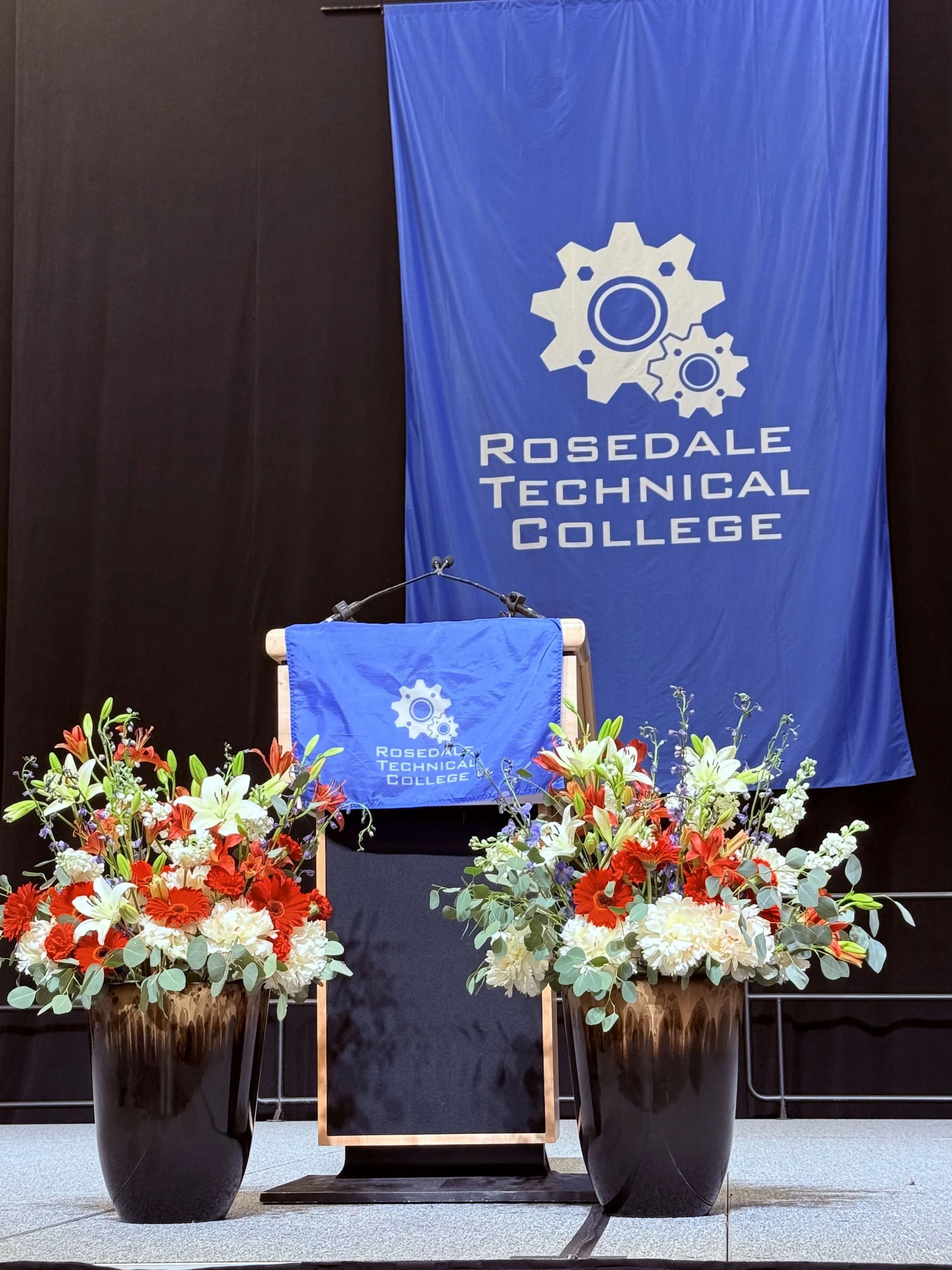 Rosedale Technical College stage with blue banner displaying gear logos, podium with a blue cloth, and two large flower arrangements with white, red, and orange flowers.