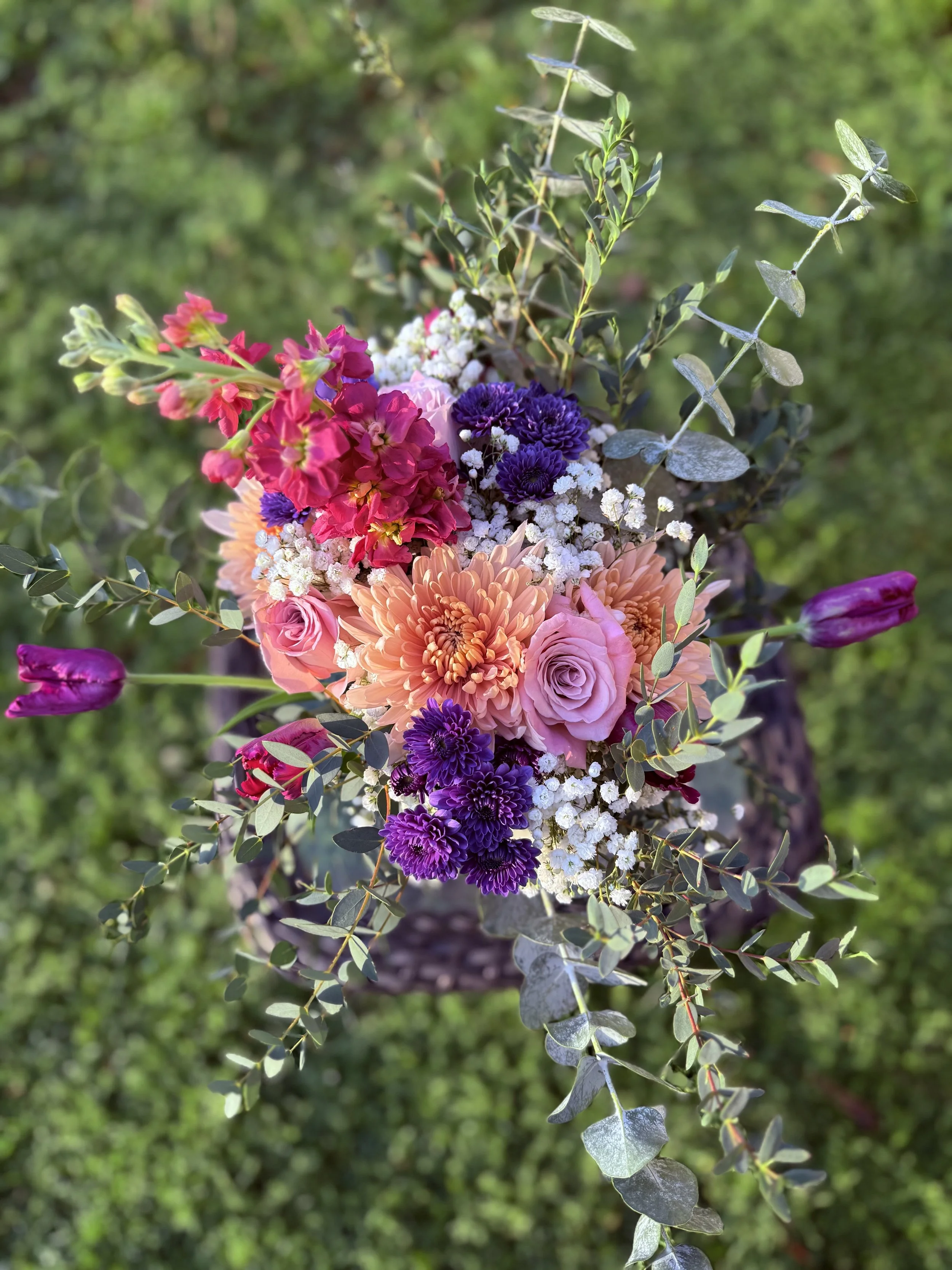 A vibrant bouquet of mixed flowers including pink roses, purple chrysanthemums, orange dahlias, red and pink snapdragons, and white baby's breath, surrounded by green eucalyptus leaves, set against a blurred green background.