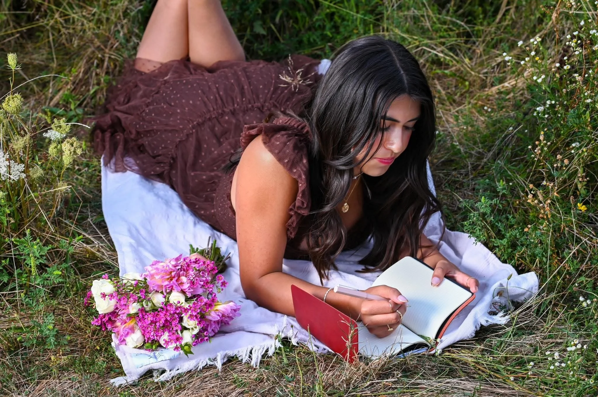 A young woman with long dark hair lying on a white blanket outdoors, writing in a notebook with a pink cover, surrounded by grass and wildflowers, with a bouquet of pink, white, and purple flowers nearby.