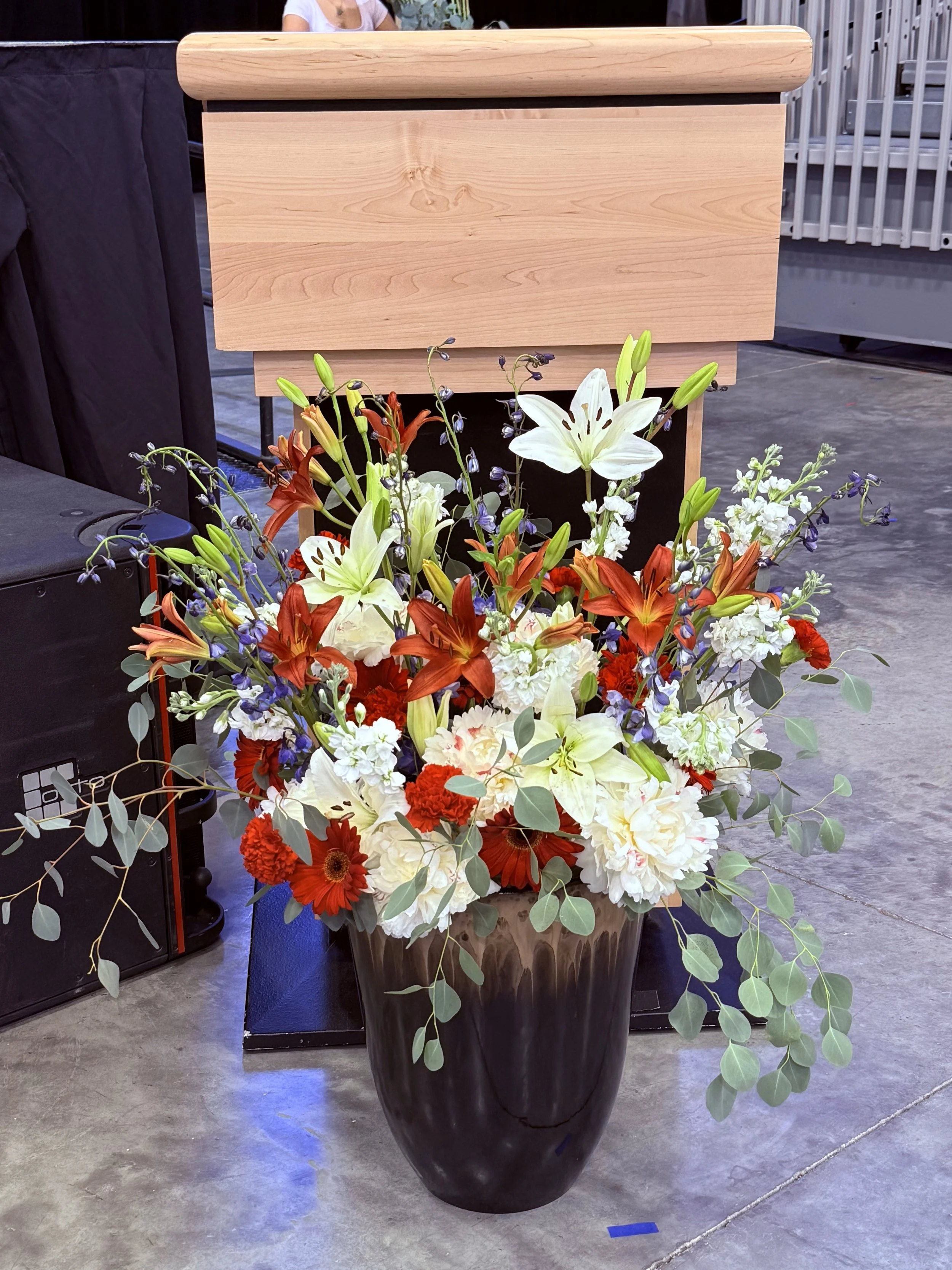 A large black vase filled with a colorful arrangement of white, red, orange, and purple flowers, including lilies, carnations, and other blooms, placed on a black pedestal on a concrete floor.